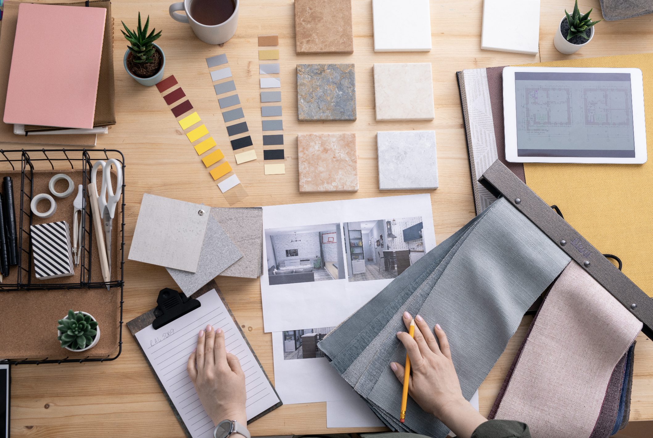 A person working on interior design or renovation plans at a desk. They are holding a color swatch book in one hand and a pencil in the other. The desk has various sample tiles, color palettes, a tablet displaying a floor plan, a printed interior picture, and some plants. There are also notebooks, scissors, and a wire organizer.
