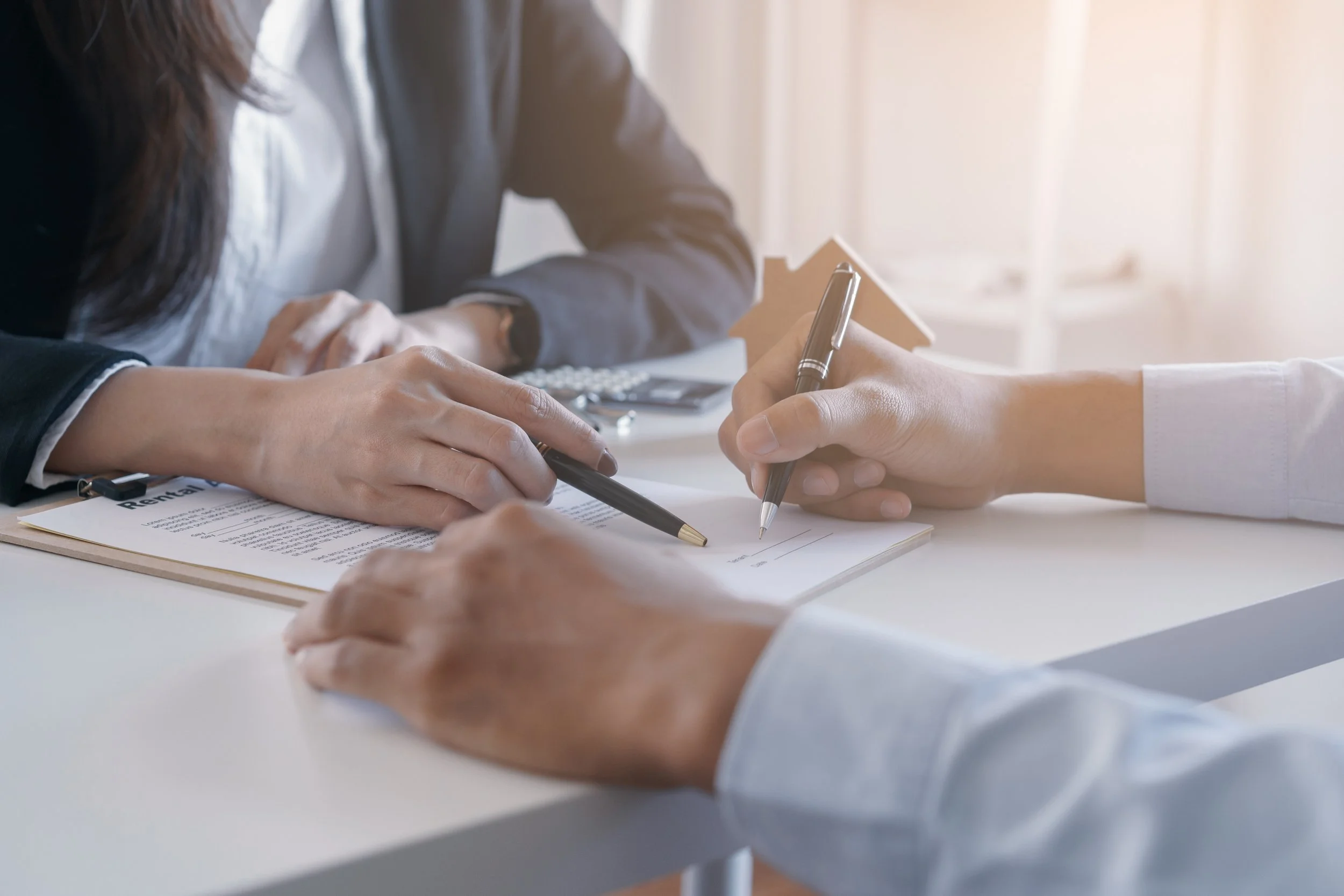 People signing a document at a desk, with one person holding a pen and others assisting, in an office setting.