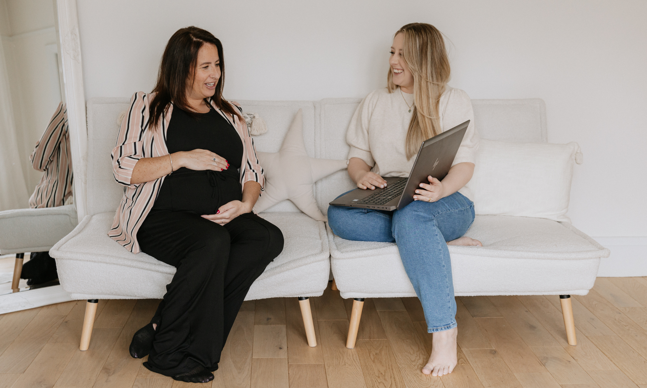 Two women sitting on a beige couch, smiling and talking with each other. One woman is holding a laptop, and they are in a brightly lit room with light-colored walls and wooden flooring.