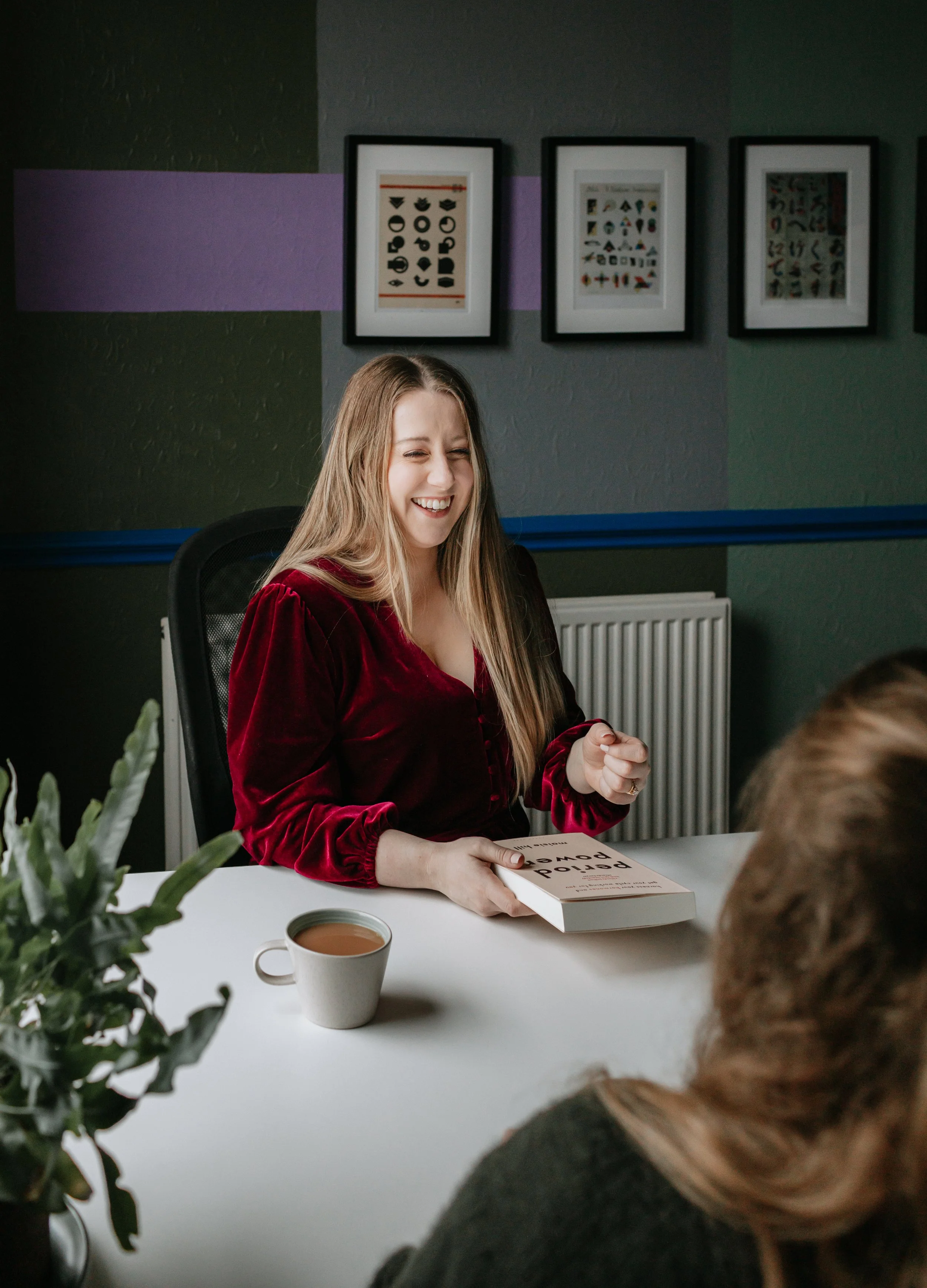 A woman sitting at a meeting table, laughing, with a book called 'Period Power' on the table, and a mug of coffee in front of her. A plant is visible in the foreground.