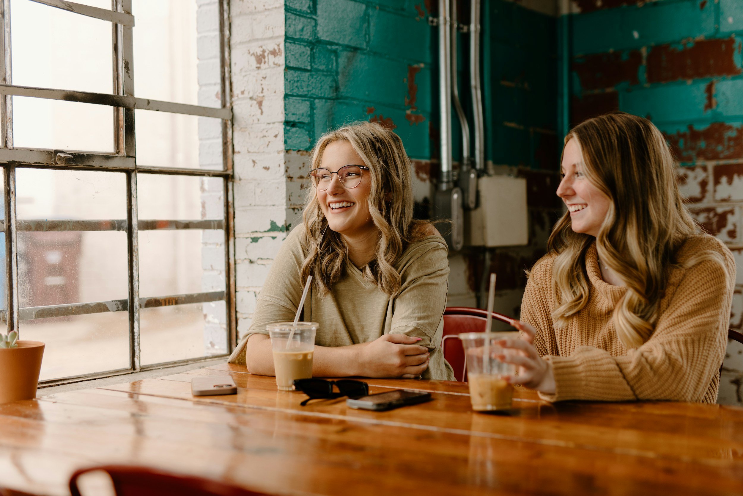 Two young women are sitting at a wooden table in a training room.  smiling and talking. They each have a drink in front of them. There are large industrial-style windows and a brick wall painted with teal and white. One woman is wearing glasses.