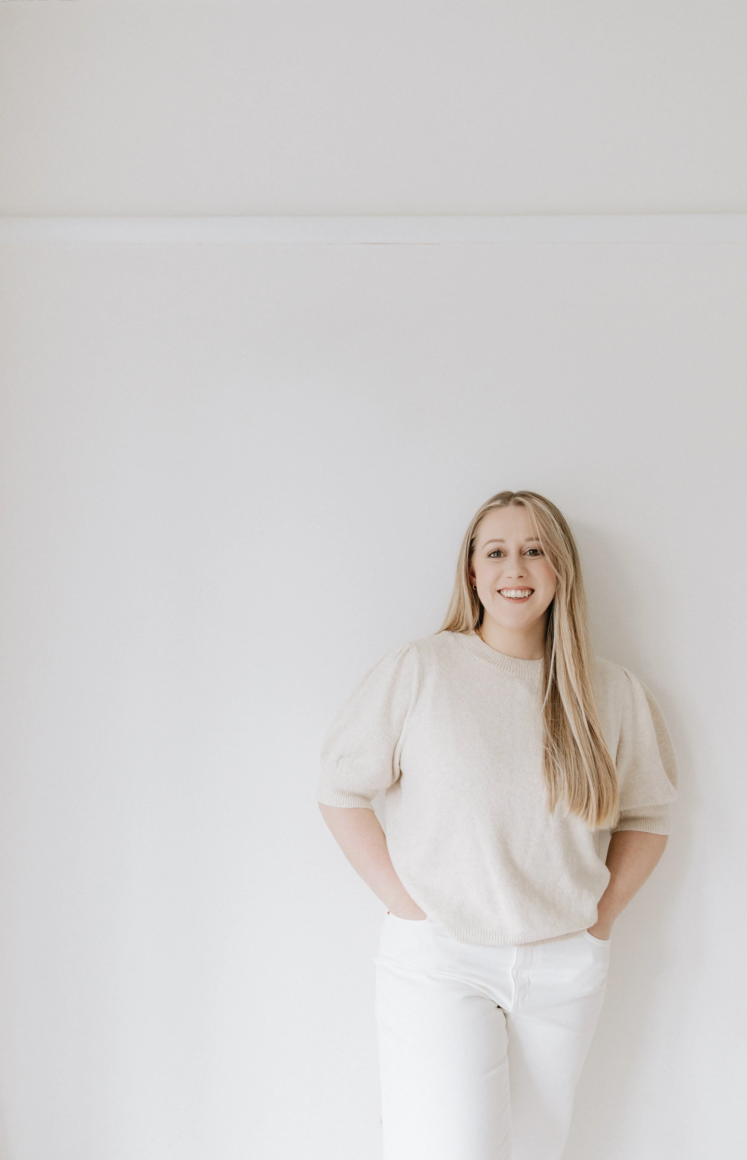 A smiling woman with long blonde hair standing against a plain white wall, wearing a beige sweater and white pants, with hands in pockets.