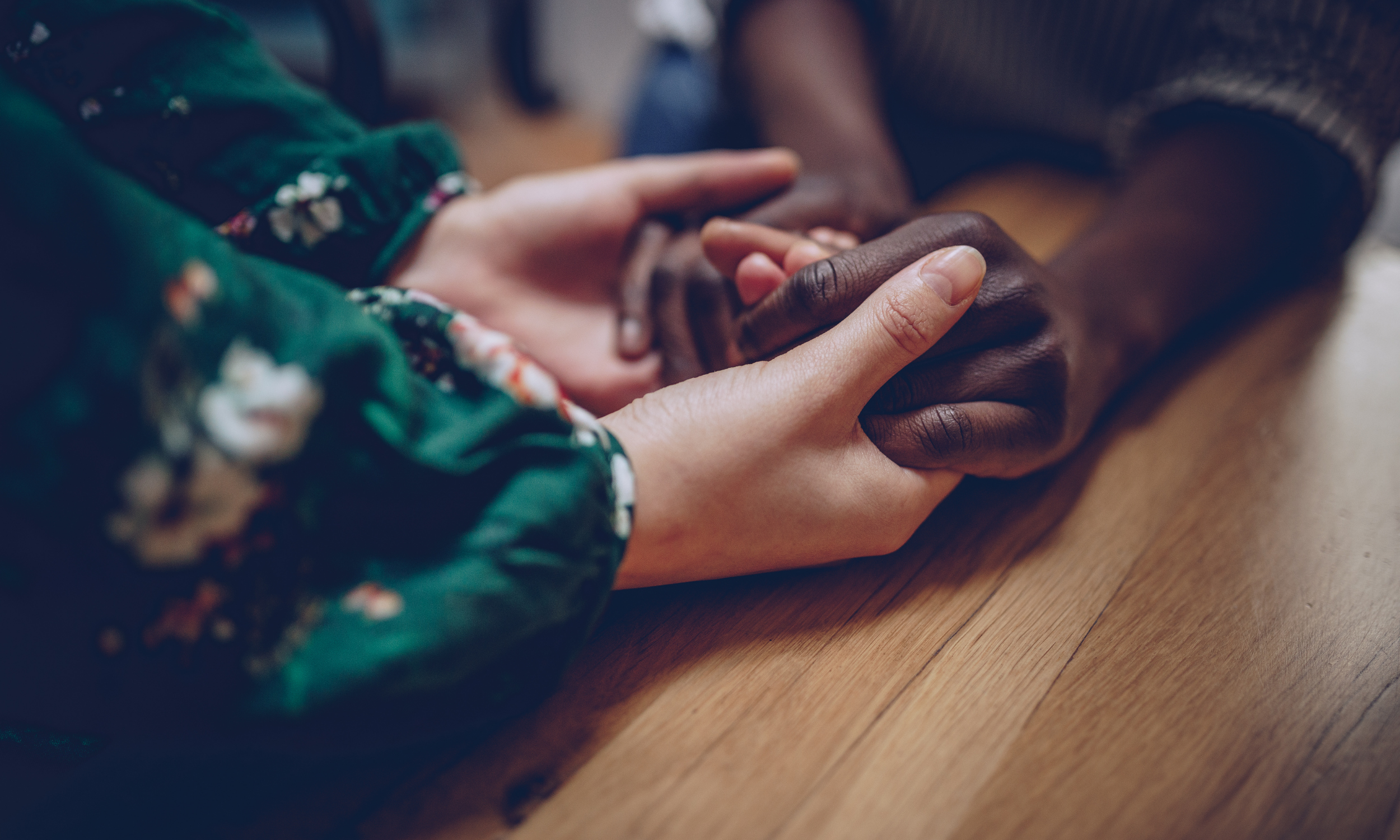 Close-up of two hands holding each other on a wooden surface, one hand with dark skin and the other with light skin, partially covered by a green floral sleeve.