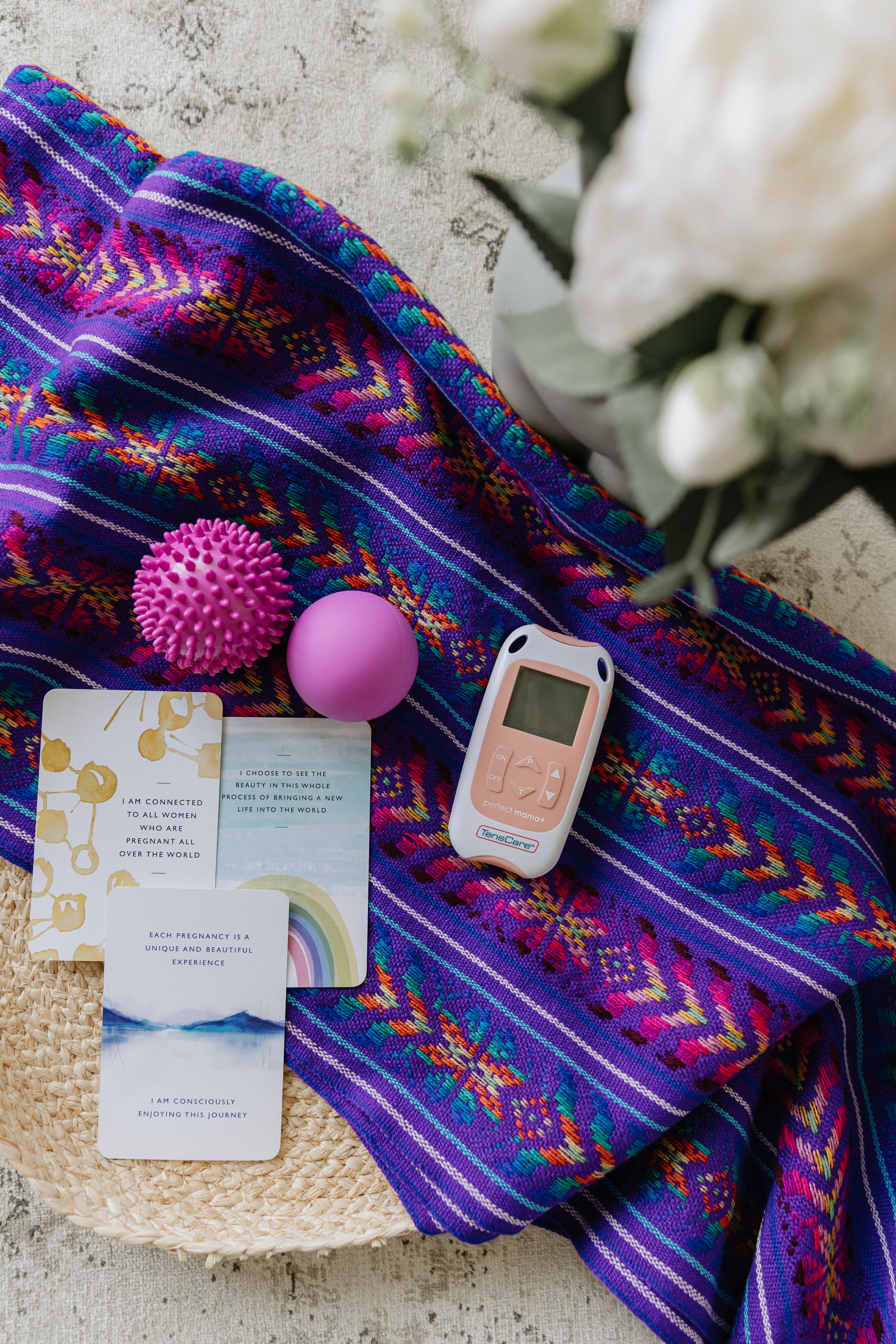 Colorful woven textile rebozo with a pink spiky massage ball, a purple ball, a tens machine, and several affirmationcards with motivational quotes, placed on a woven round surface with a beige textured background.