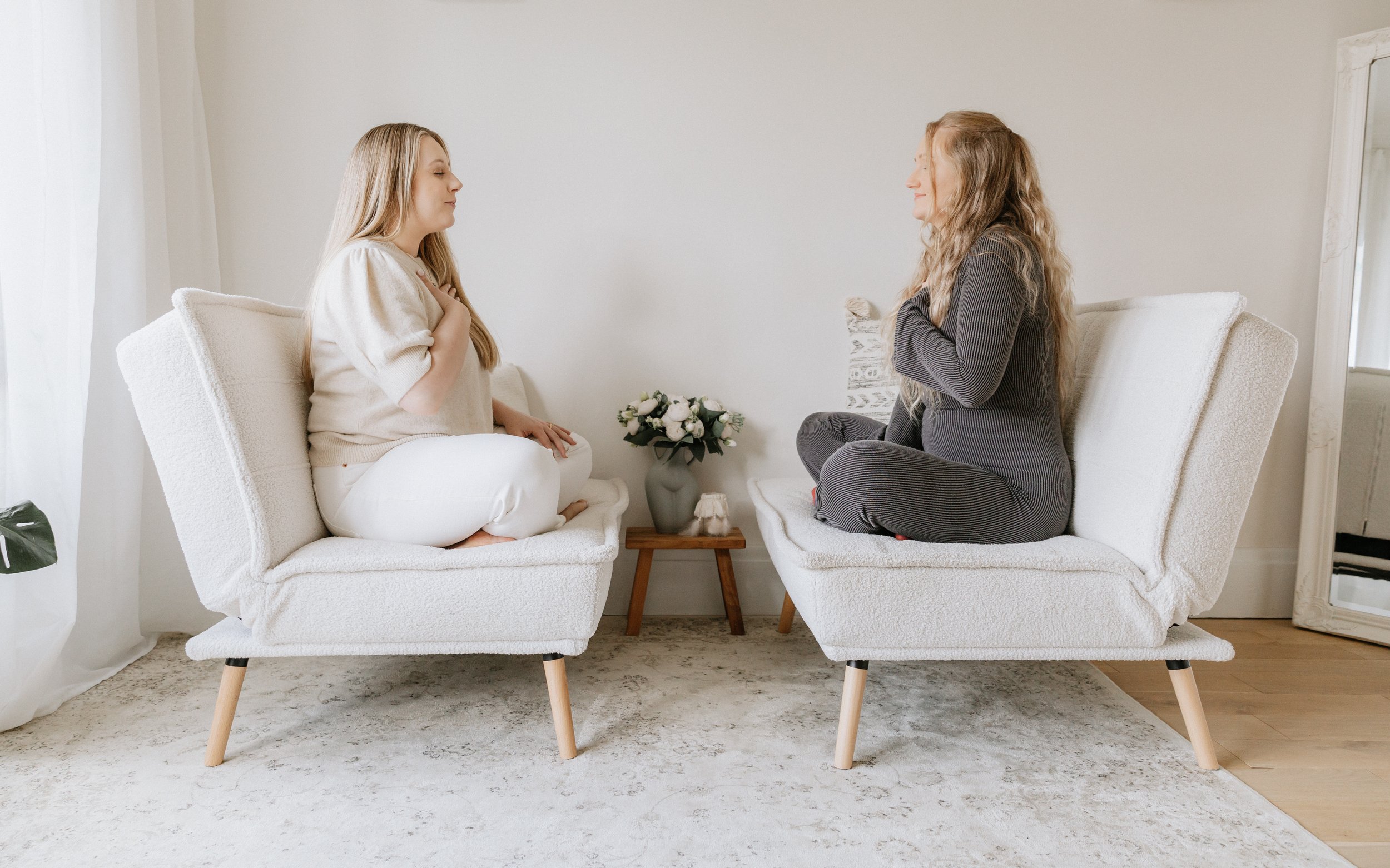 Two women sitting on separate white chairs facing each other, with palms on their chests, in a minimalist room with neutral colors, a small wooden table with a bouquet of white flowers between them, and a large mirror on the wall.