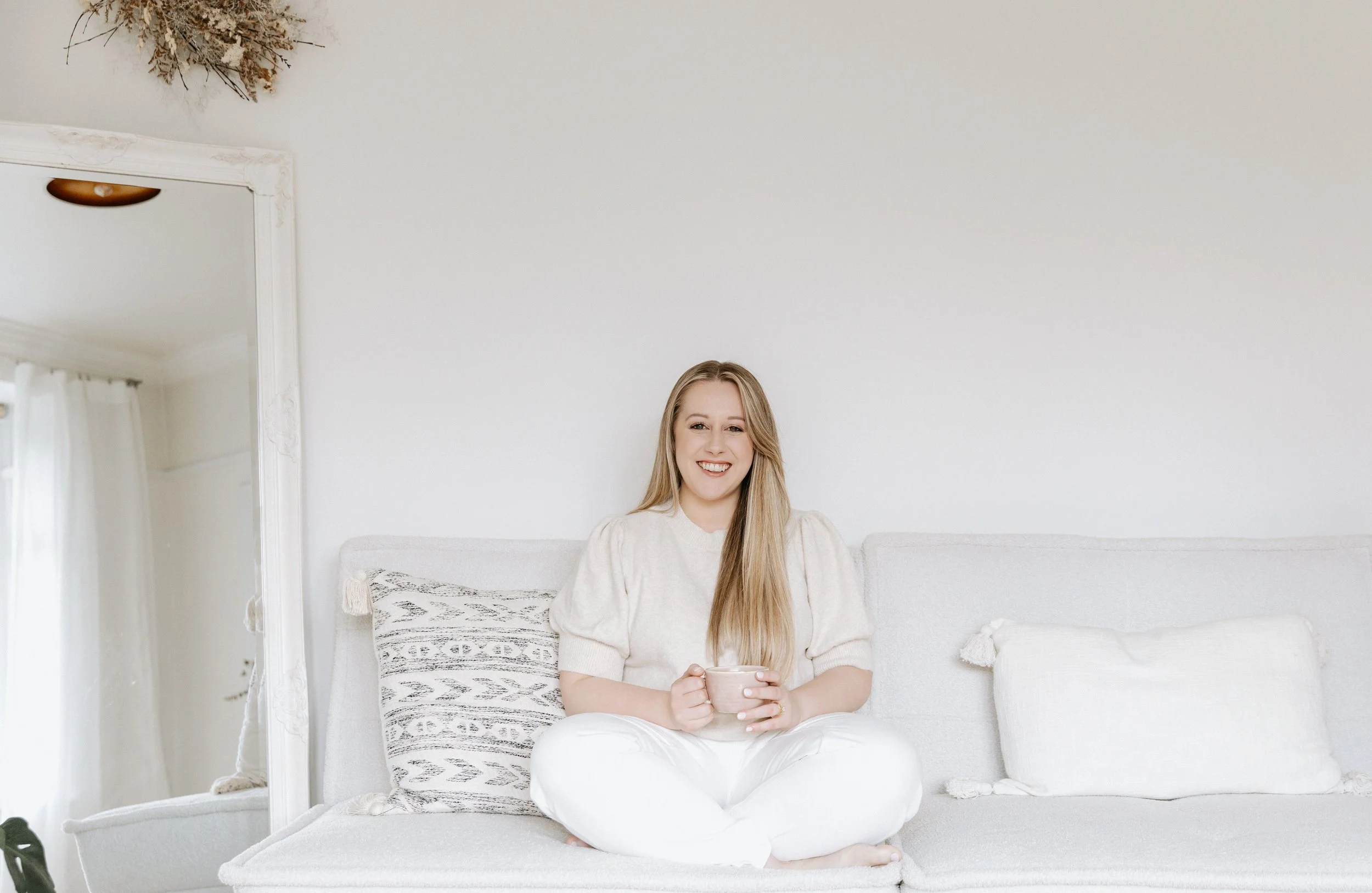 A doula with long blonde hair sitting on a white sofa holding a mug, smiling, in a bright room with white walls and curtains.