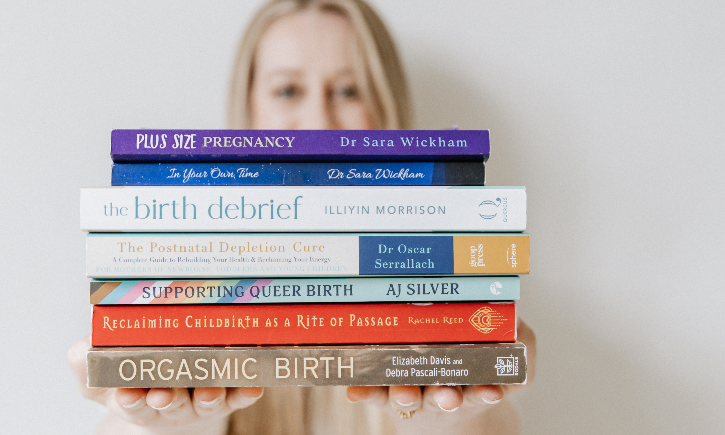 A doula holding a stack of books about pregnancy, birth, and reproductive health, with the person's face partially obscured behind the books.