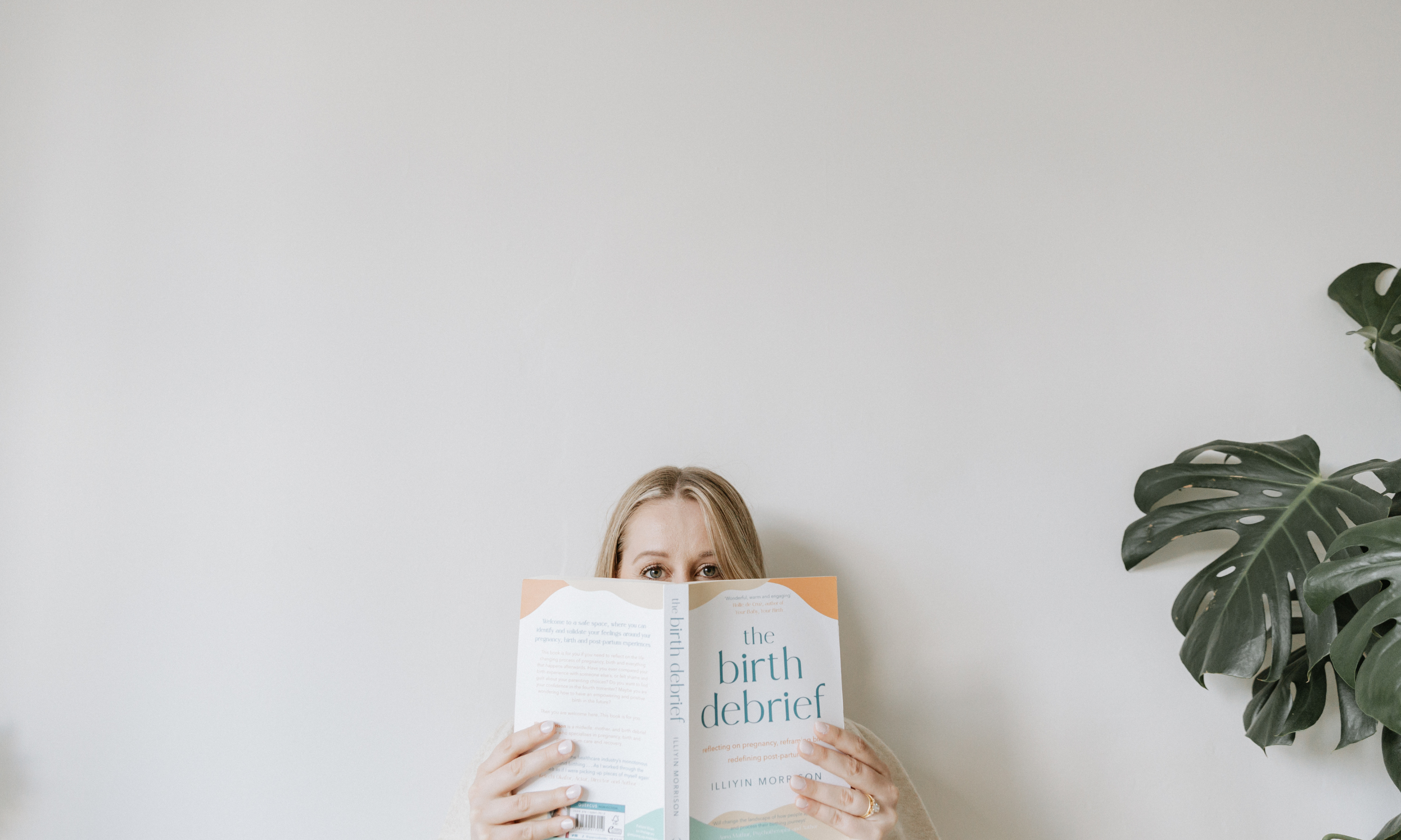 A woman with blonde hair holding a book titled 'the birth of a debrief' in front of her face, with only her eyes and forehead visible, standing near a white wall with a large green plant on the right side.