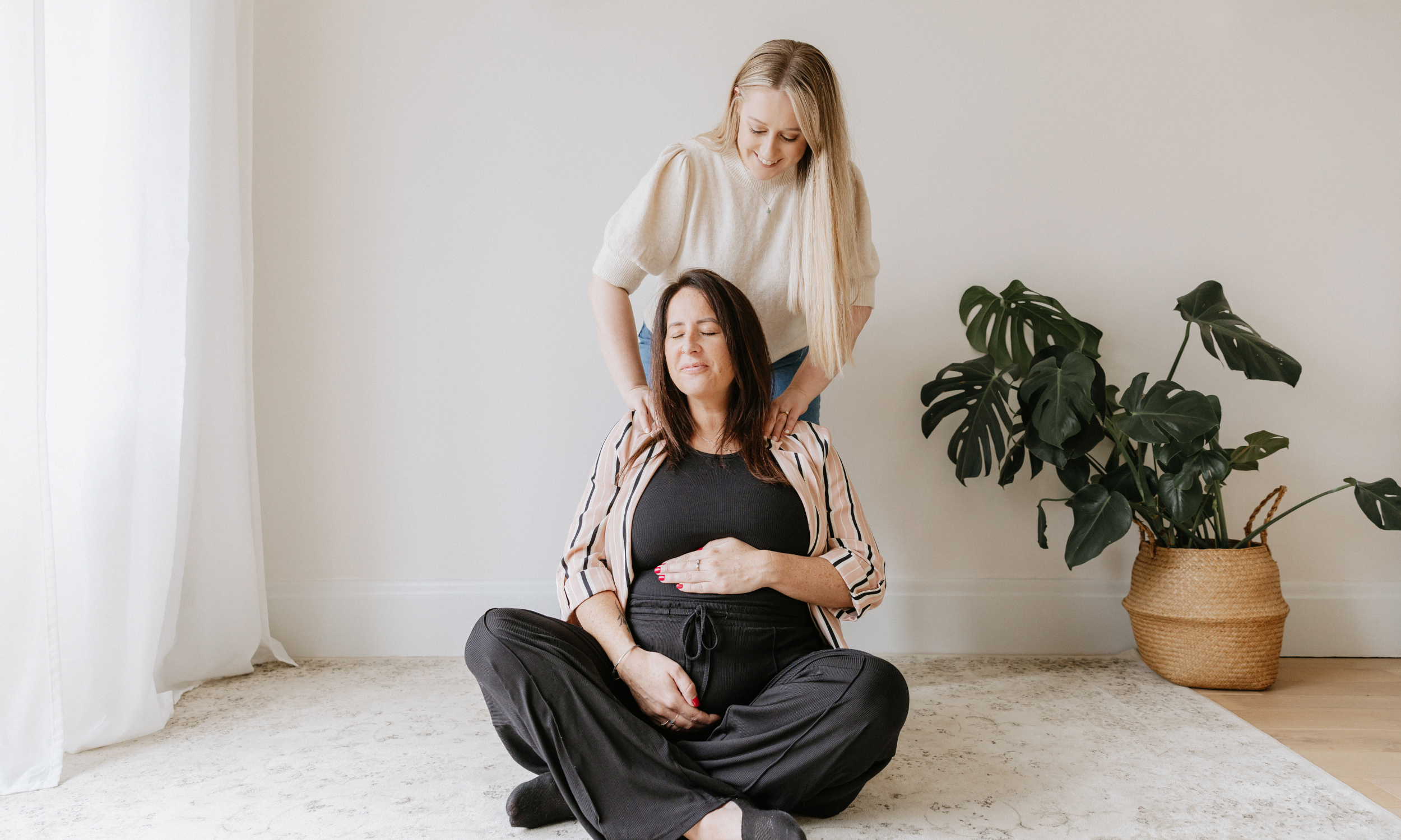Two women, one sitting cross-legged on a light-colored carpet with her eyes closed and hands on her pregnant stomach, and the other, standing behind her gently touching her shoulders, in a bright, minimalistic room with a large green potted plant.