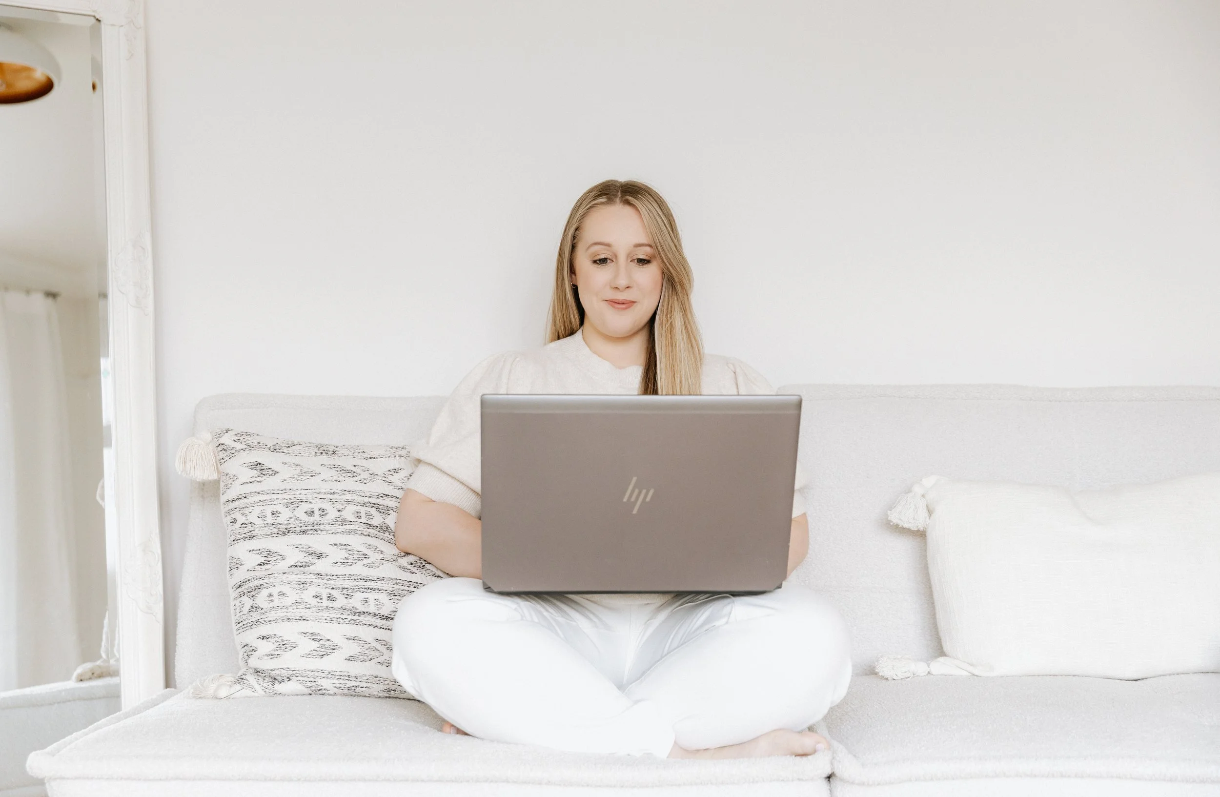 A woman with long blonde hair sitting cross-legged on a white couch, working on a silver HP laptop, in a bright room with minimal decor and white walls.
