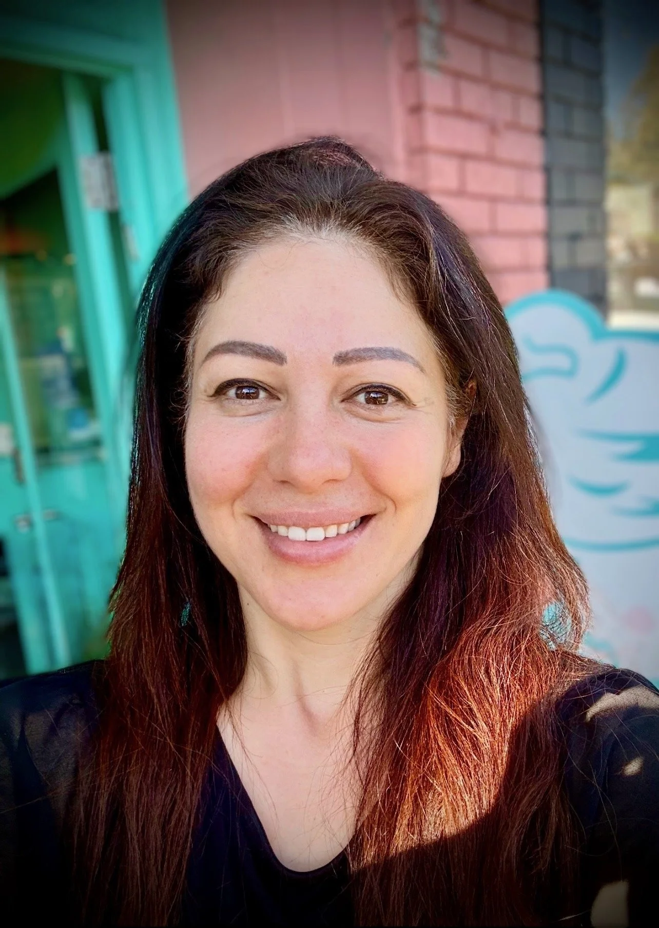 A woman with long, reddish-brown hair smiling outdoors against a background of brick wall and a teal-colored window.