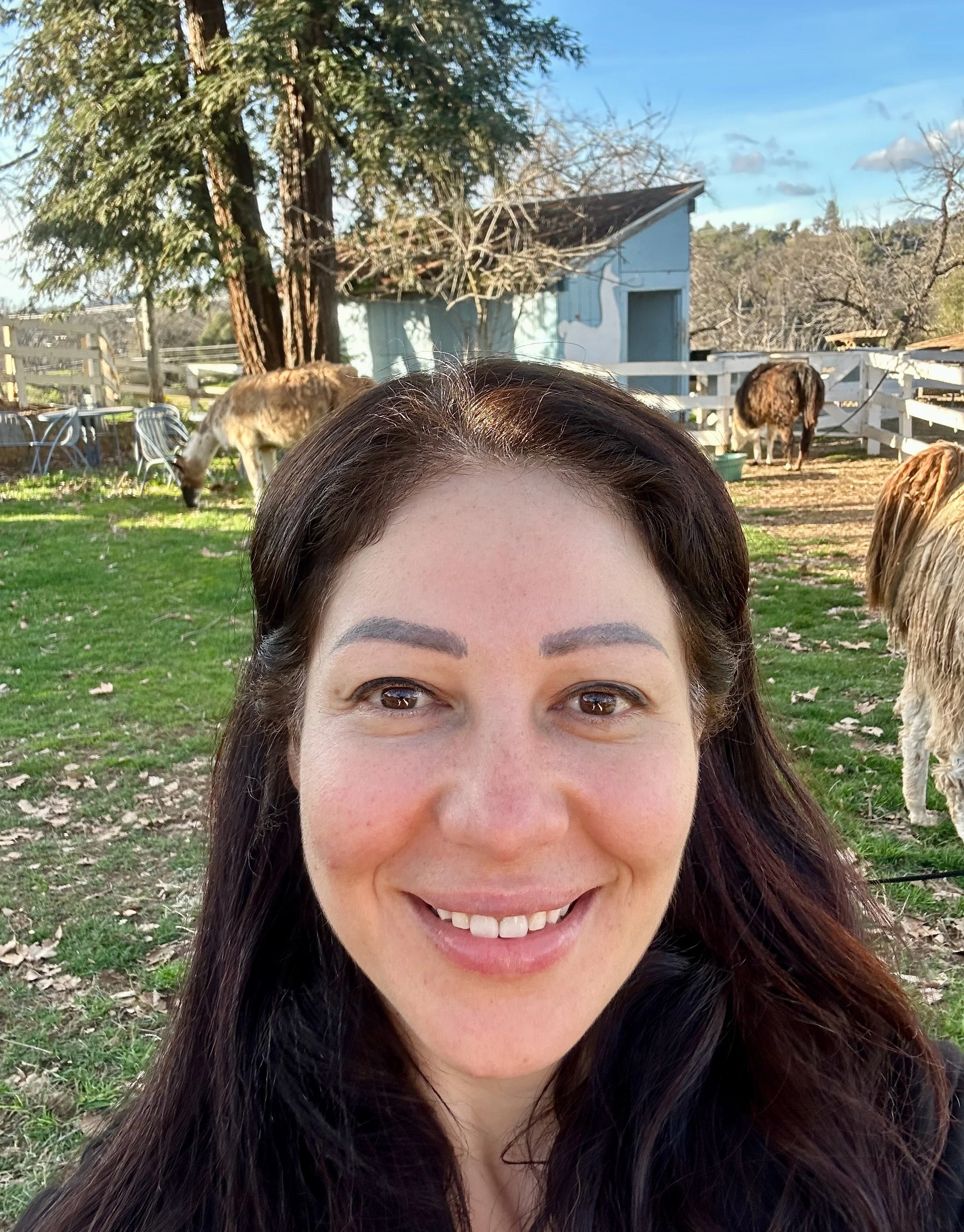 A woman with dark brown hair smiling at the camera outdoors, with guinea pigs, a small blue building, trees, a white fence, and a blue sky in the background.