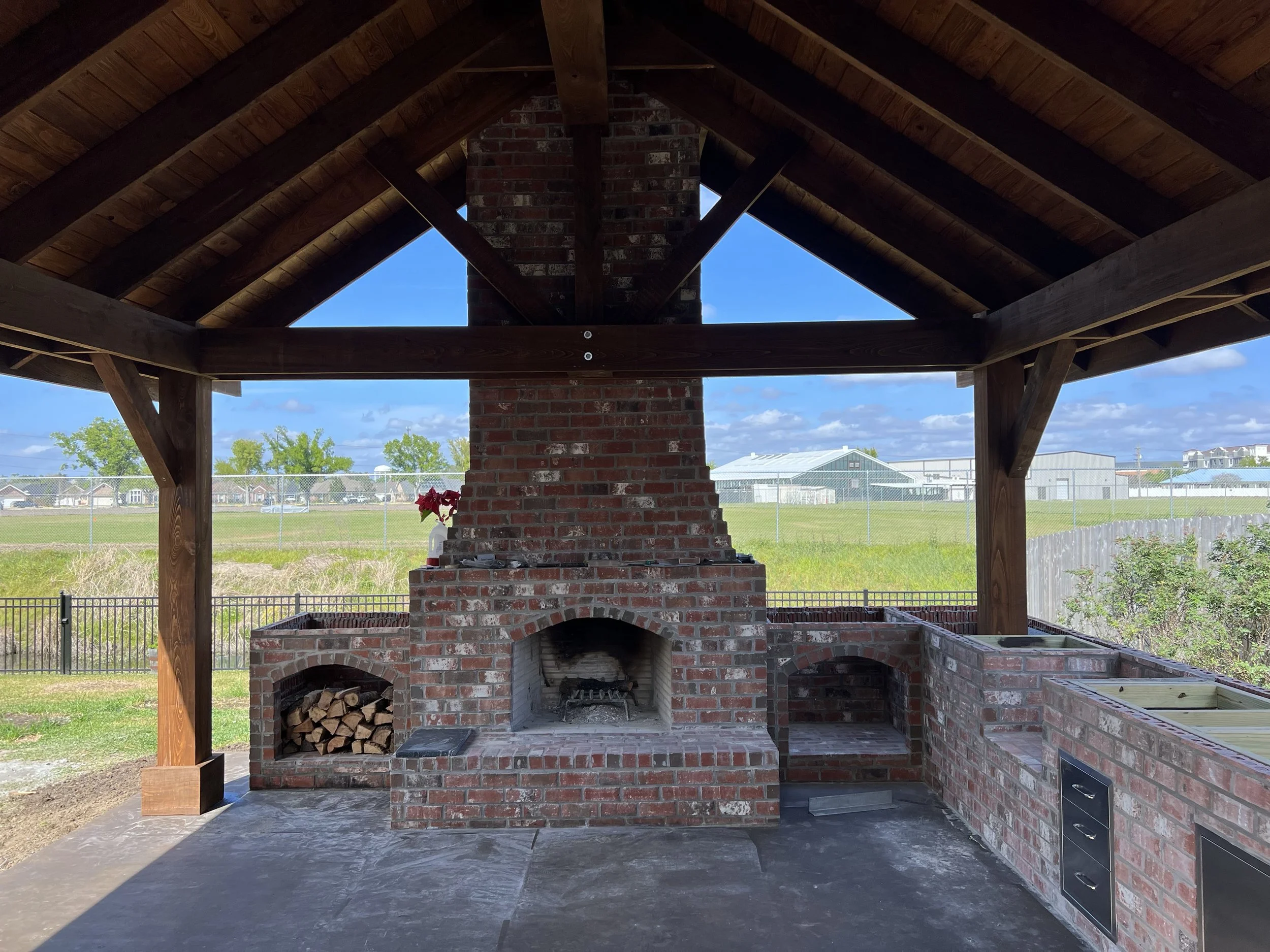 Outdoor brick and wood fireplace with a chimney, under a wooden roof structure, overlooking a grassy field and residential area.