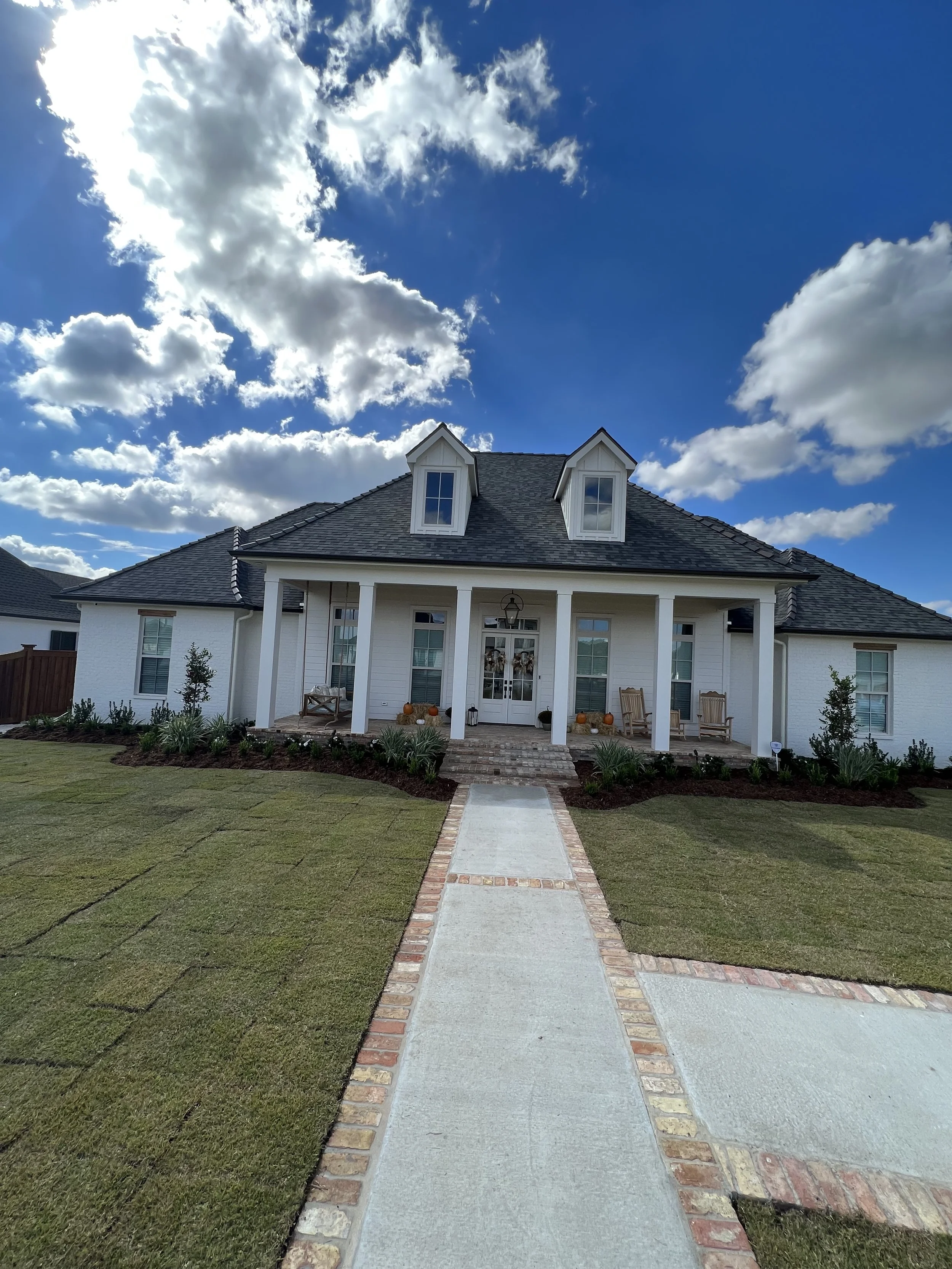 Front view of a white house with a dark gray roof, a covered porch with white columns, and a landscaped yard with a brick-bordered walkway leading to the front door. The sky is partly cloudy with white clouds and blue sky visible.