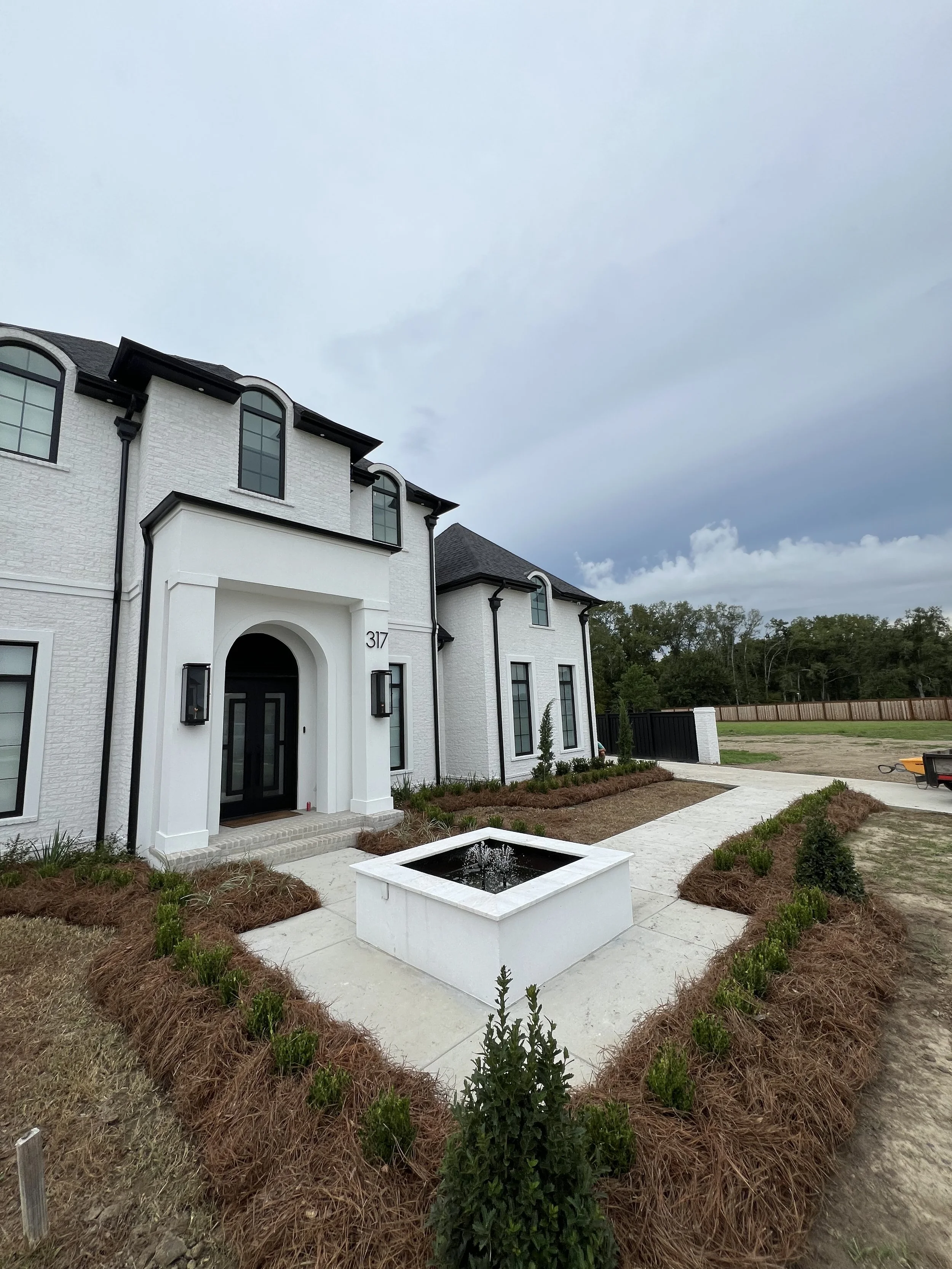 Front view of a white house with arched windows, a black front door, and a small fountain in the front yard, surrounded by landscaping with shrubs and mulch, under a cloudy sky.