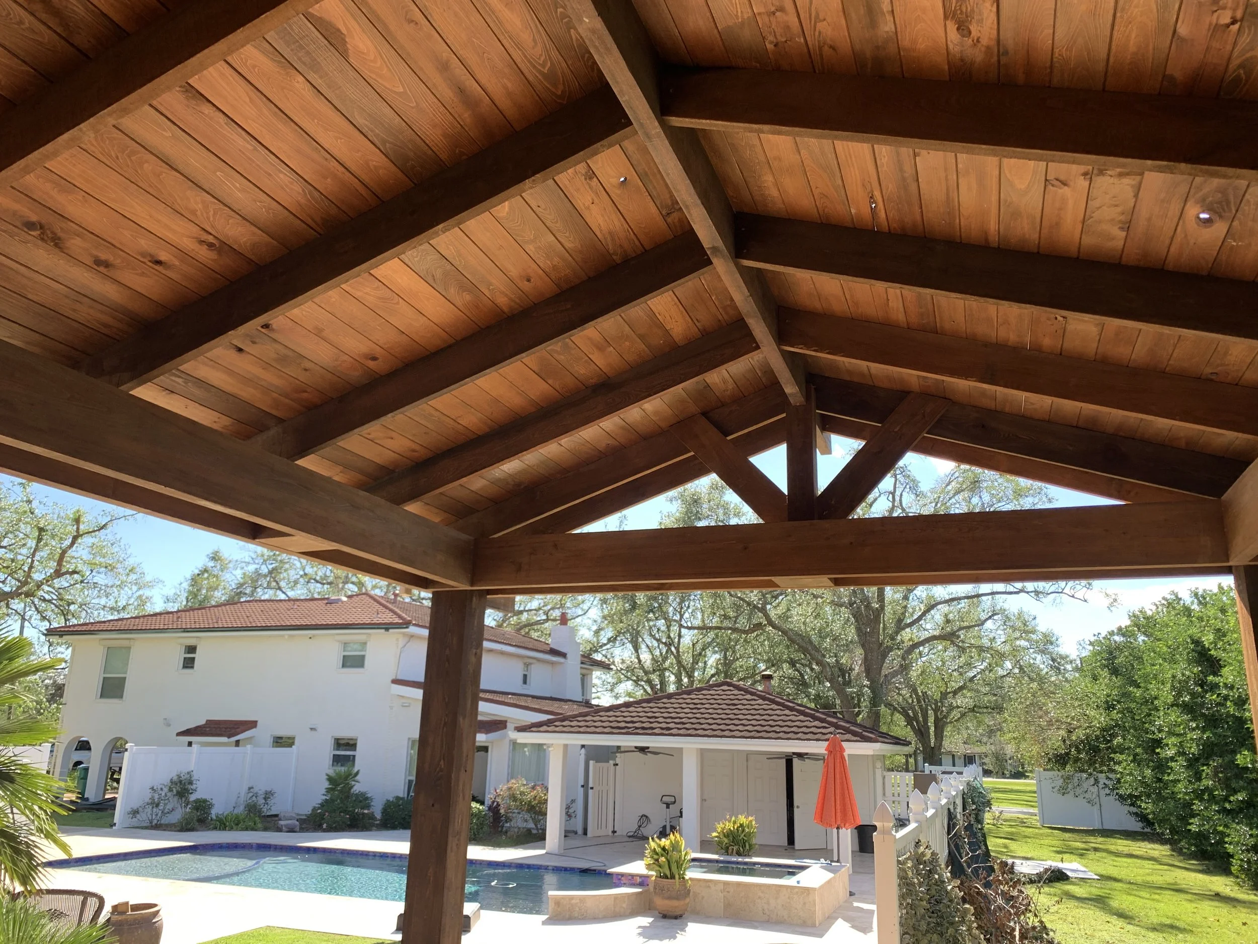 Backyard view featuring a wooden pergola with a sloped roof, a swimming pool, a white house with multiple windows, a backyard fence, some trees, patio furniture, and an orange umbrella.