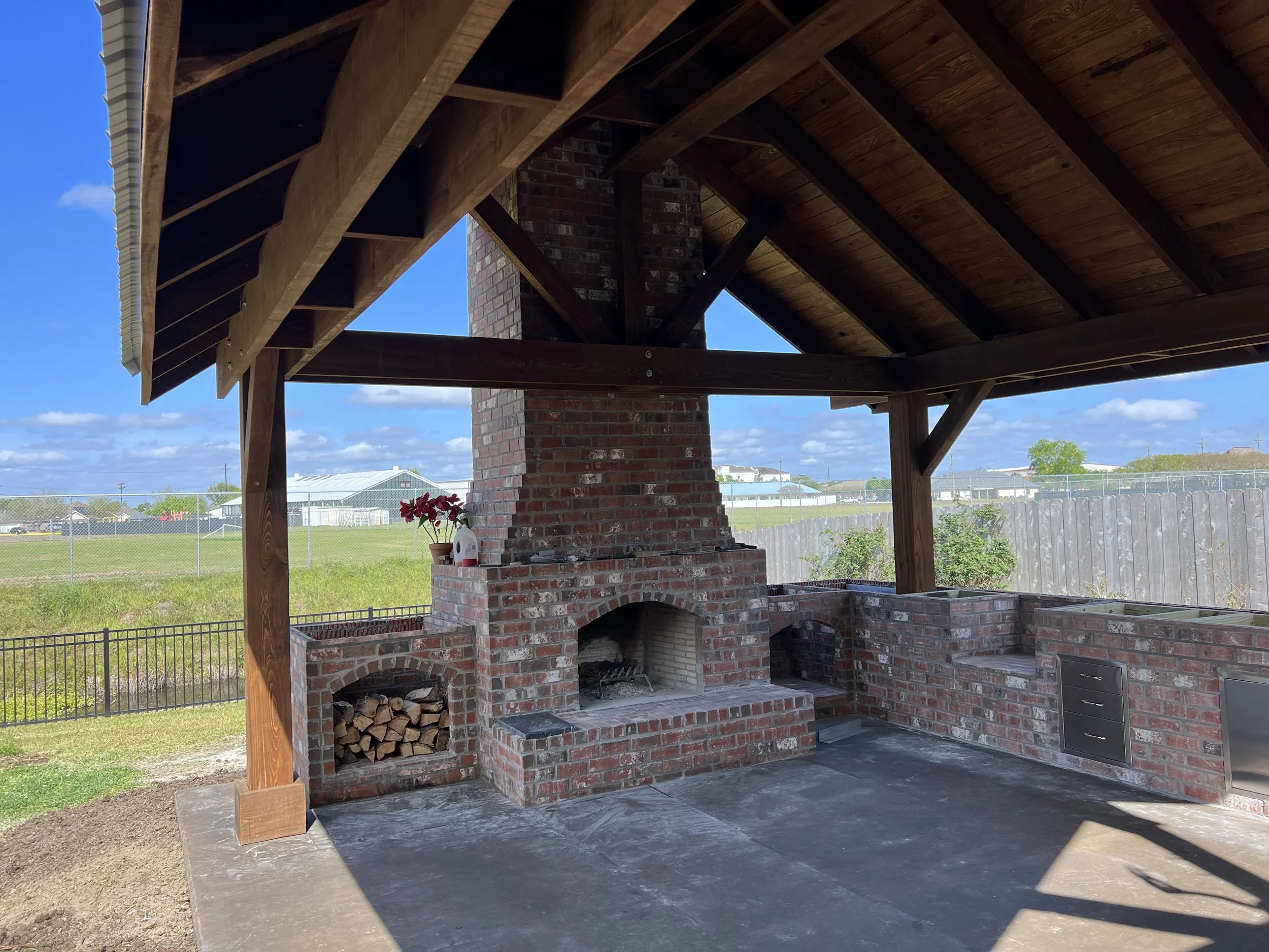An outdoor brick fireplace and barbecue area with a chimney, wood storage, and a brick countertop, under a wooden pergola with a clear blue sky in the background.
