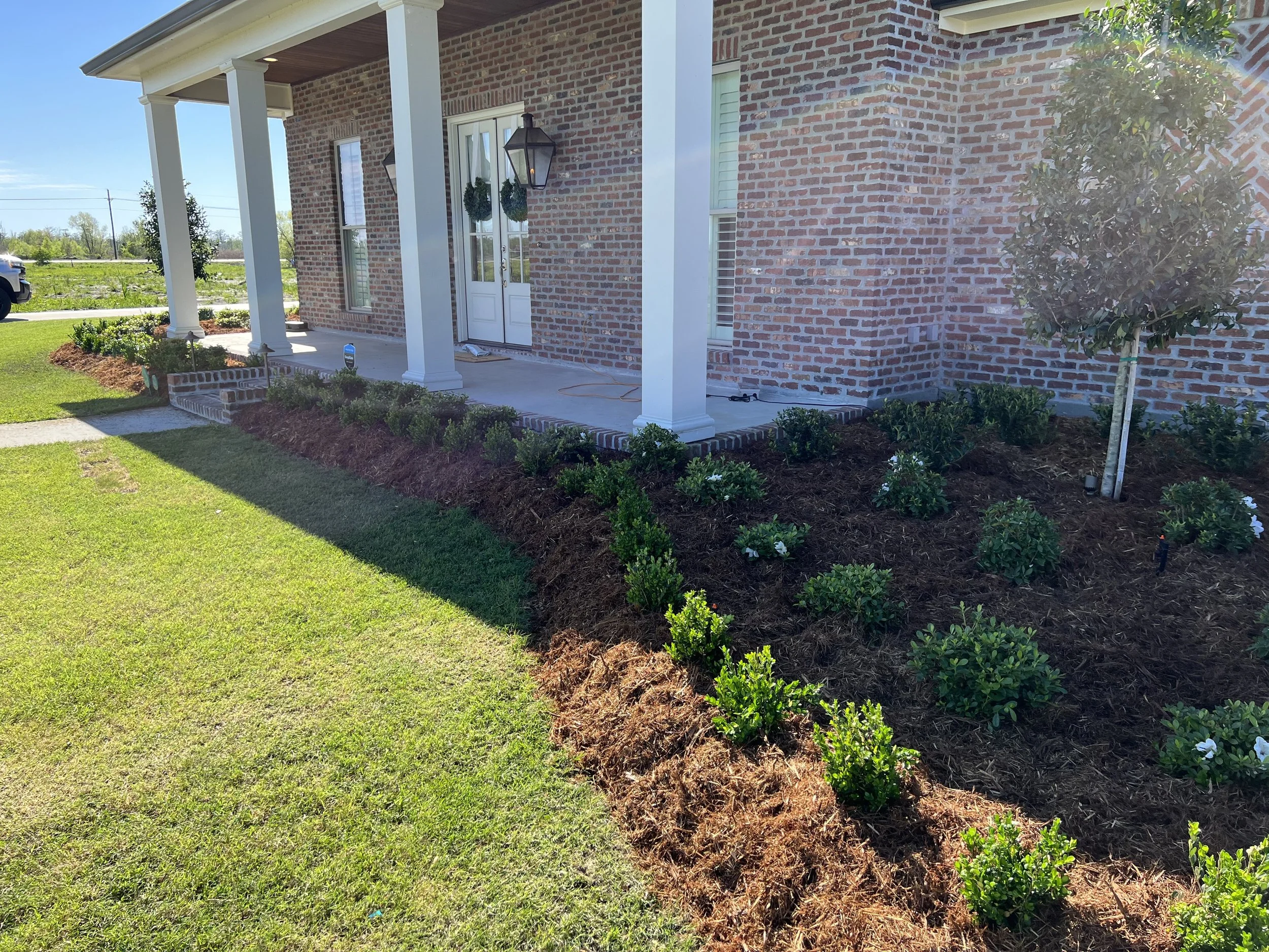 Front porch of a brick house with white columns, hanging planters, and a landscaped flower bed with small bushes and white flowers.
