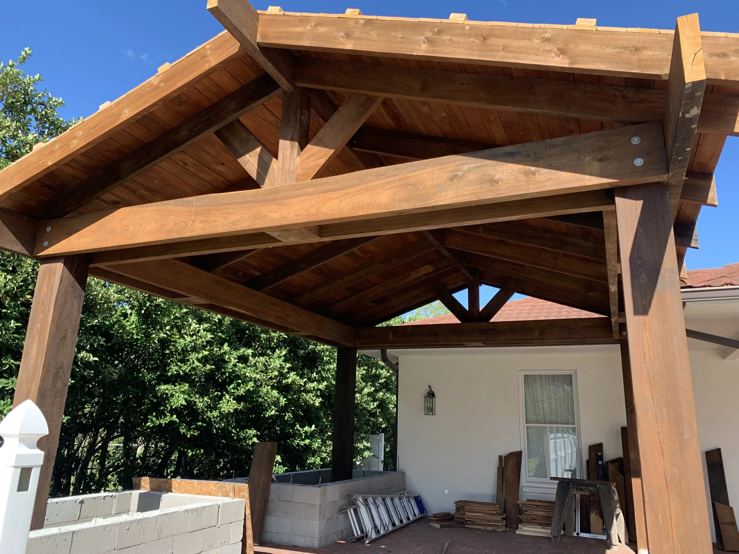 Under construction wooden patio cover with support beams, stored construction materials, and a ladder, next to a house with a window and outdoor light fixture, and lush green trees in the background.