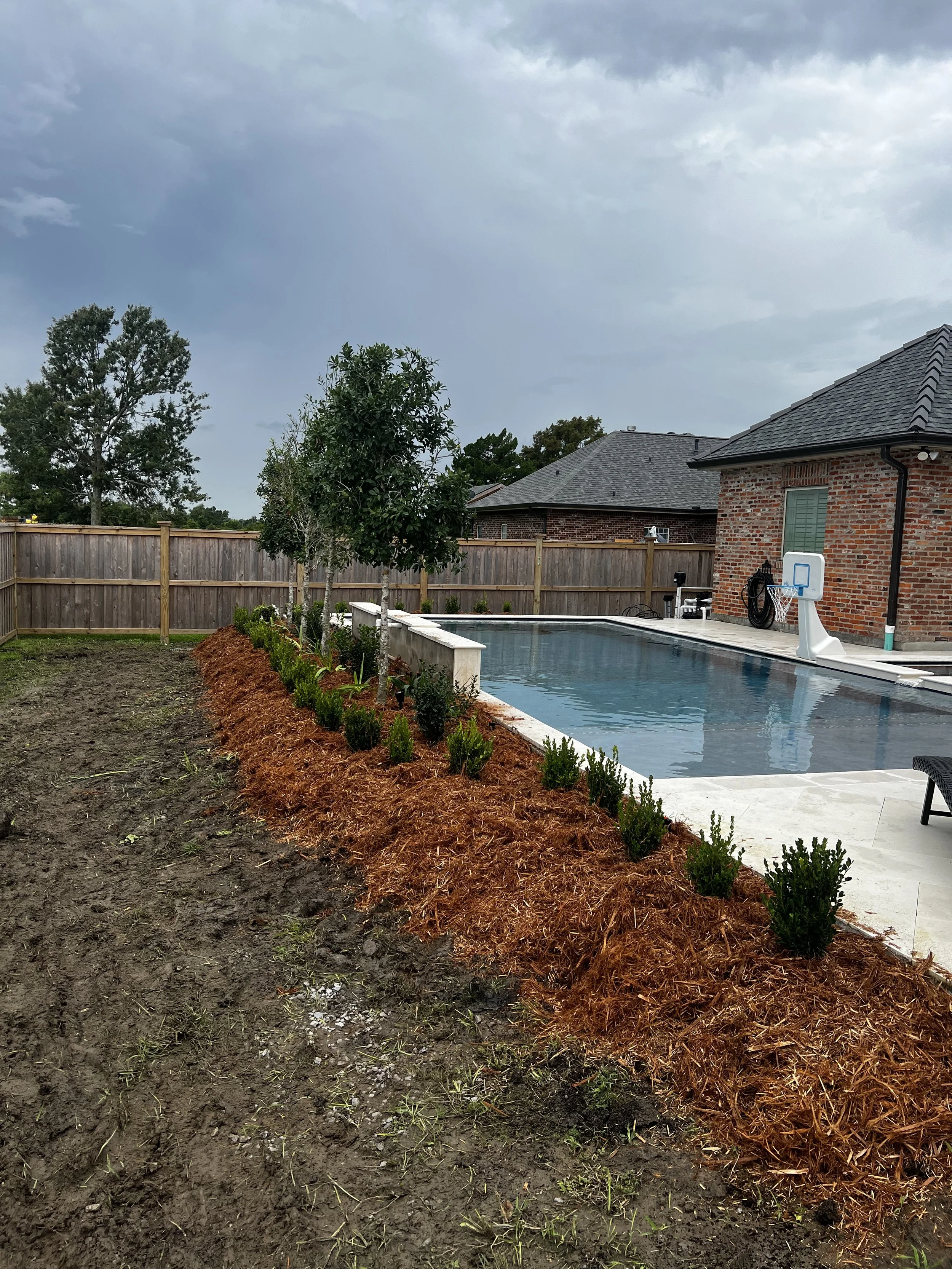Backyard with an in-ground pool, a brick house, a basketball hoop, trees, a wooden fence, and a cloudy sky.