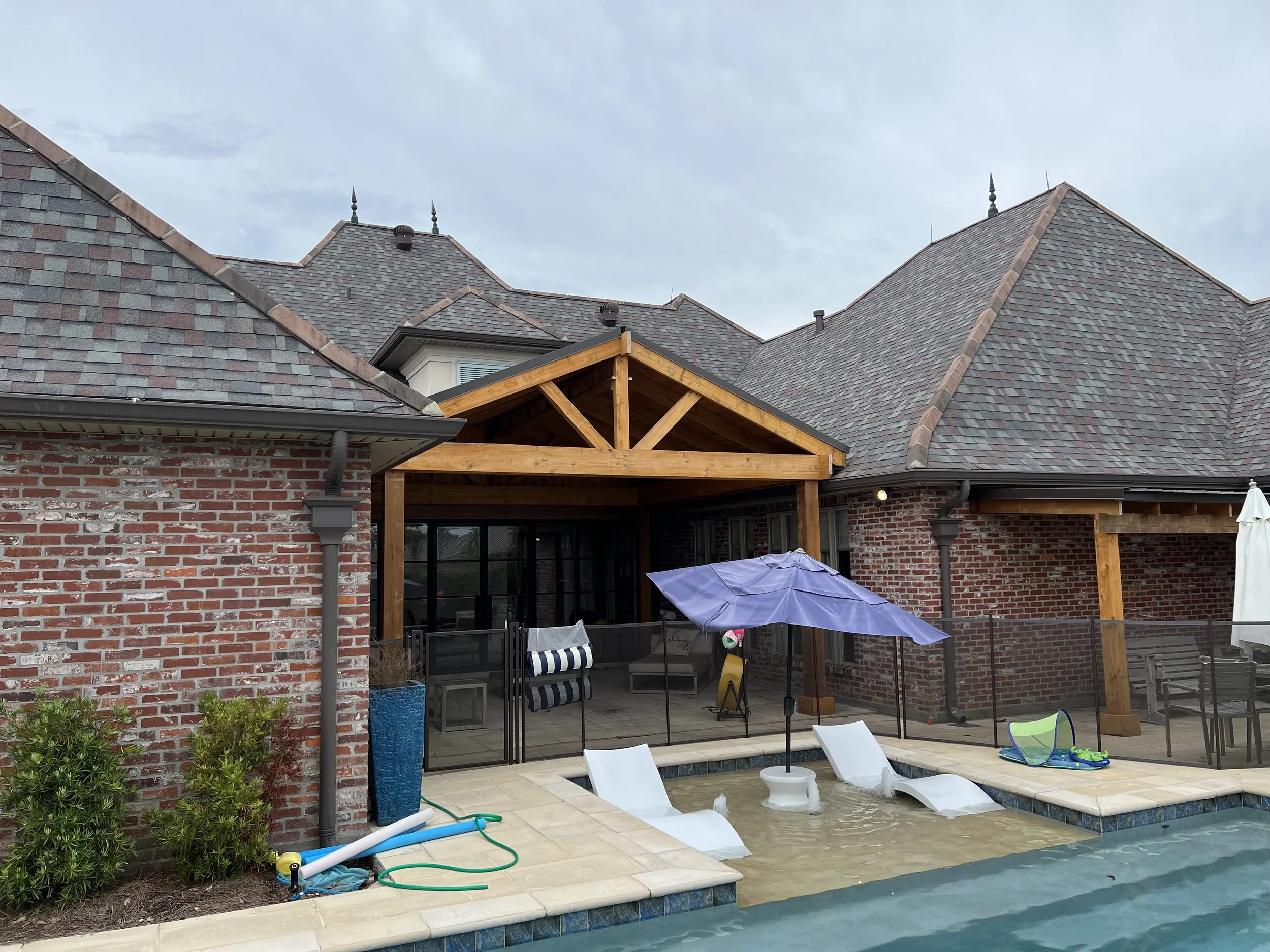 A backyard scene with a swimming pool and lounge chairs, surrounded by a brick house with a roof, some patio furniture, umbrellas, and construction materials.