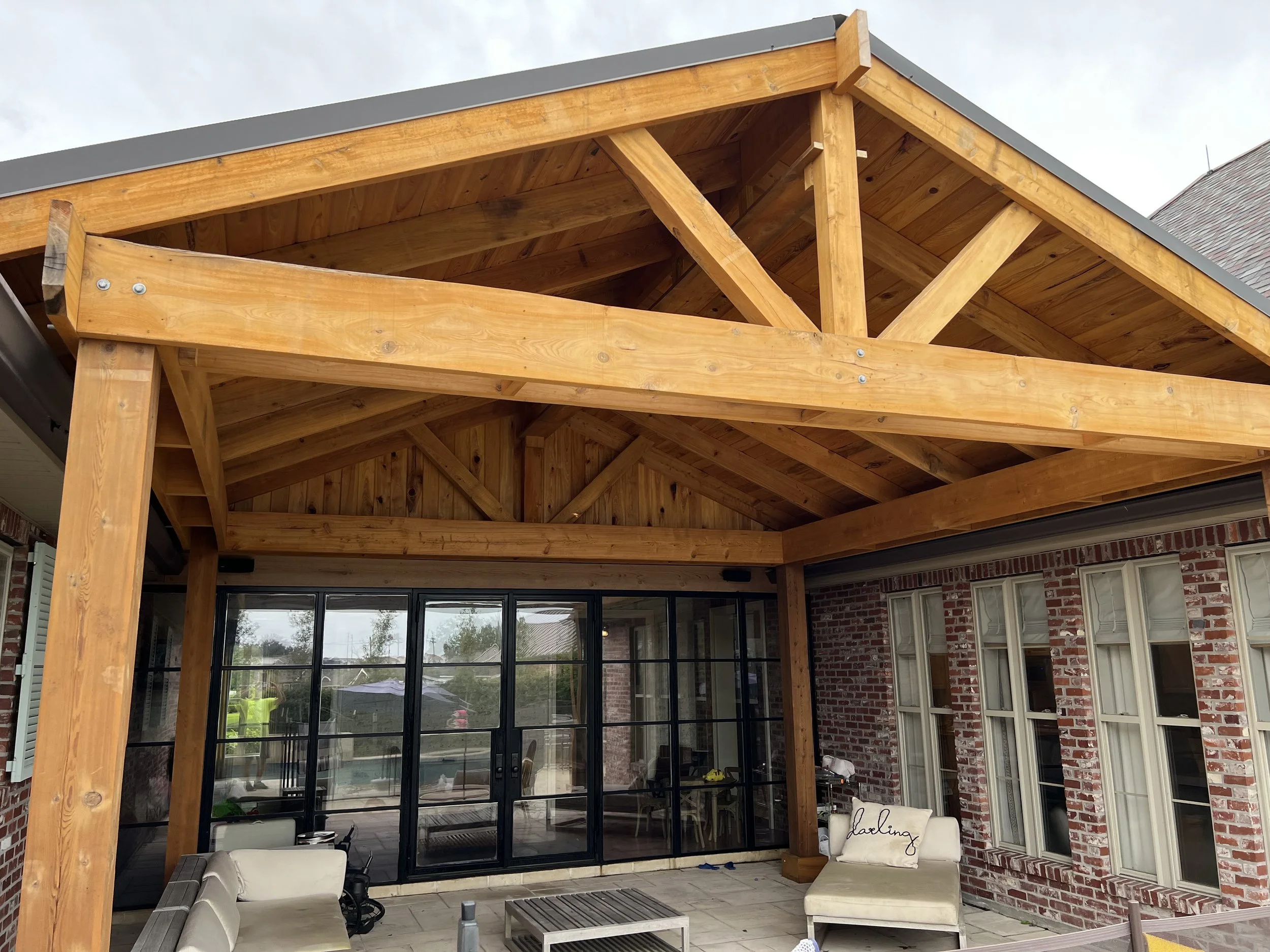 Backyard patio with a wooden pergola, outdoor seating with cushions, and a brick house with large windows.