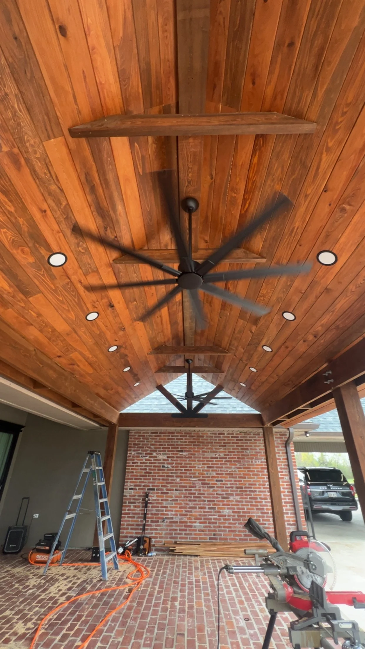 This is an outdoor covered patio with a wooden ceiling, ceiling fans, and brick wall. Tools and equipment are visible, indicating ongoing construction or renovation.