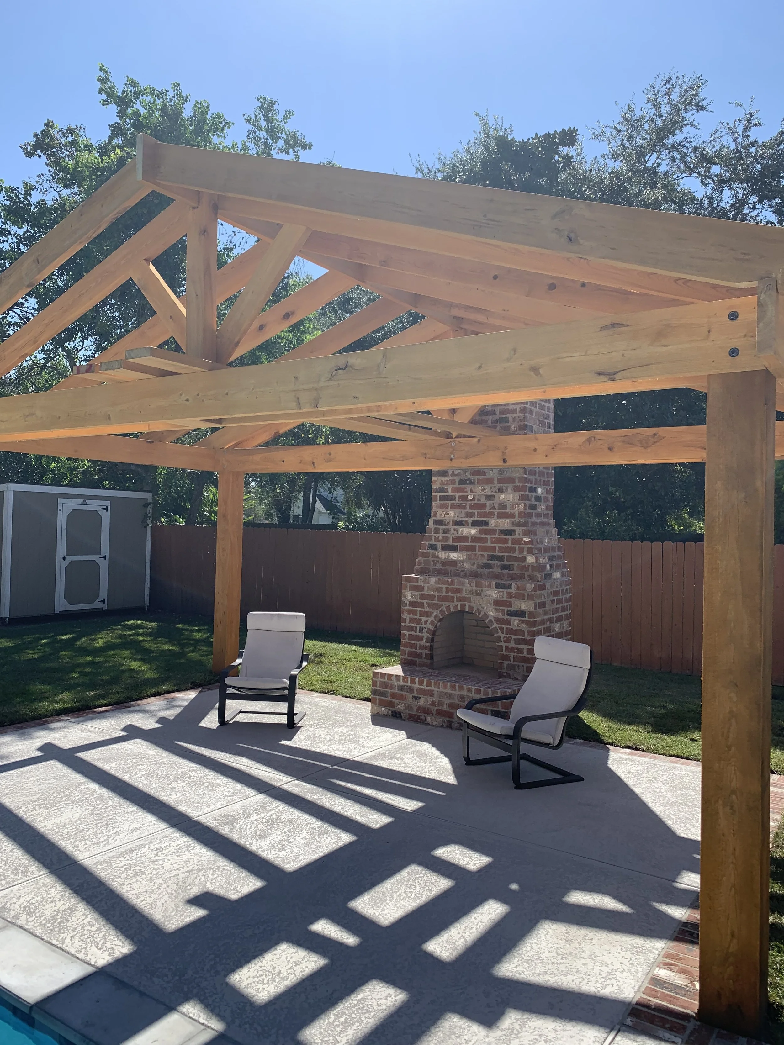 A backyard patio with two white chairs near a brick fireplace, under a wooden pergola casting striped shadows on the concrete floor.