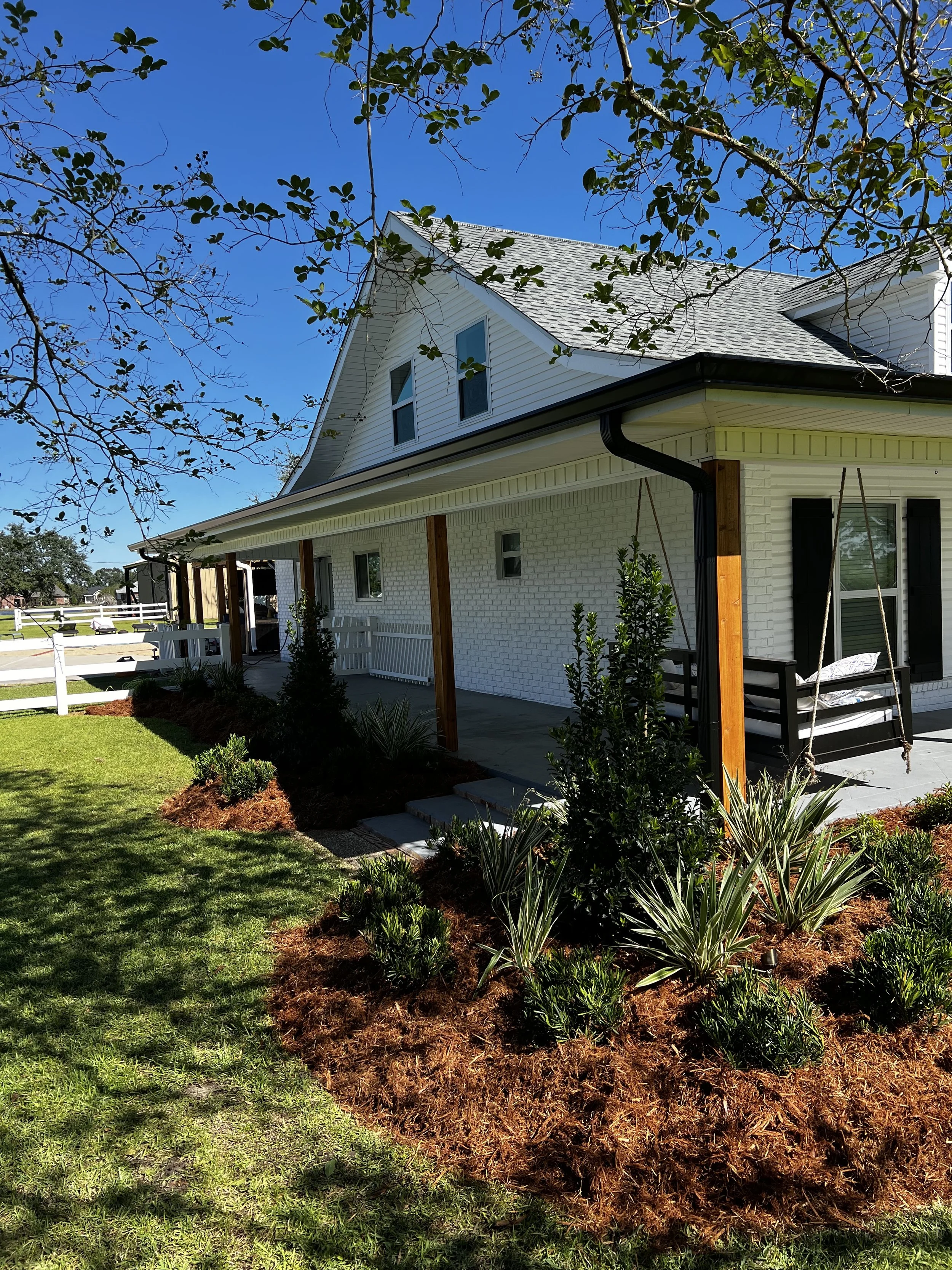 A white house with black shutters and a front porch with a swing, surrounded by landscaped garden beds with shrubs and plants, on a sunny day with clear blue sky.