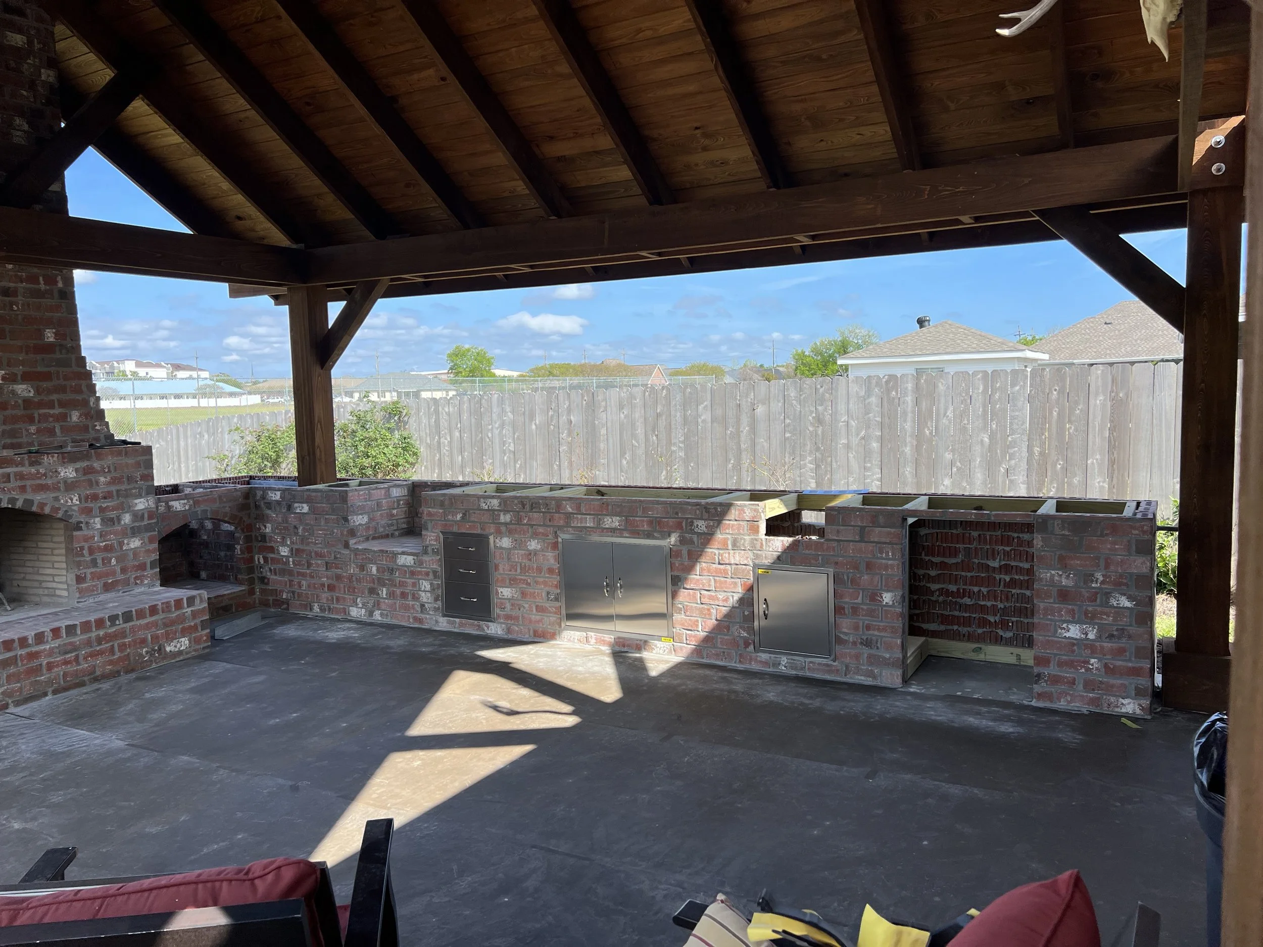 A partially built outdoor brick kitchen with a brick counter, a built-in grill, and stainless steel cabinets, under a wooden roof supported by wooden beams, with a backyard and neighboring houses in the background.