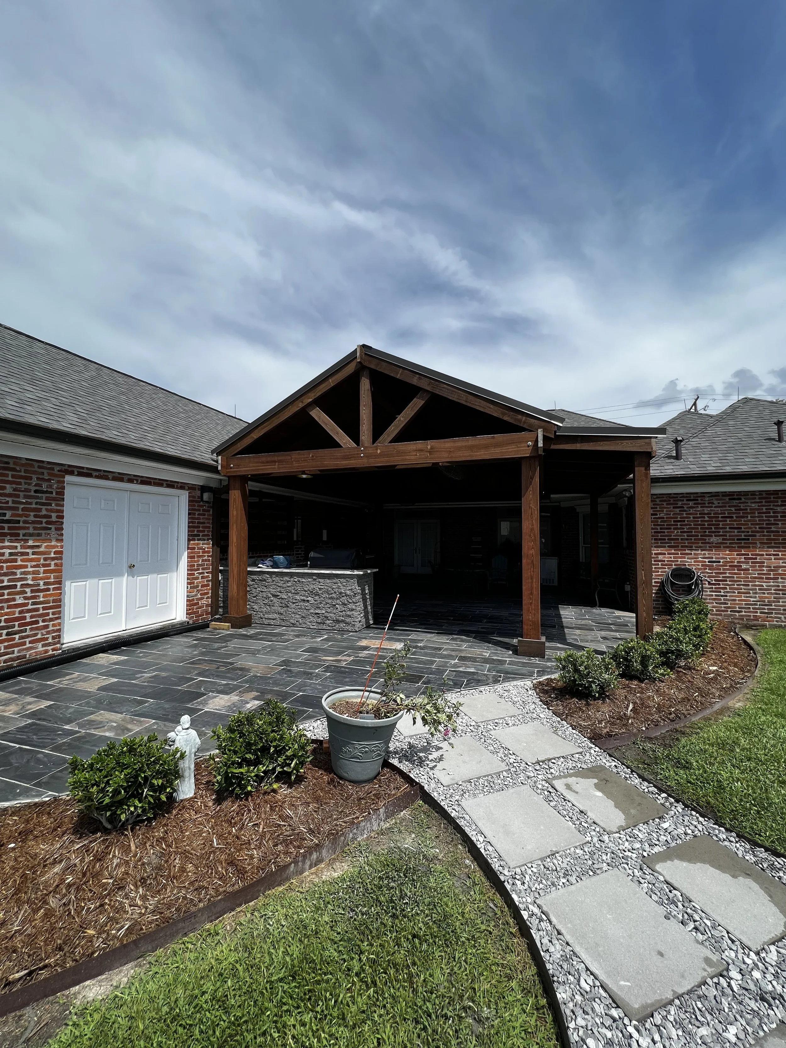 Backyard patio with stone flooring, a covered wooden pergola, landscaping with plants, and a brick house with a garage under a partly cloudy sky.