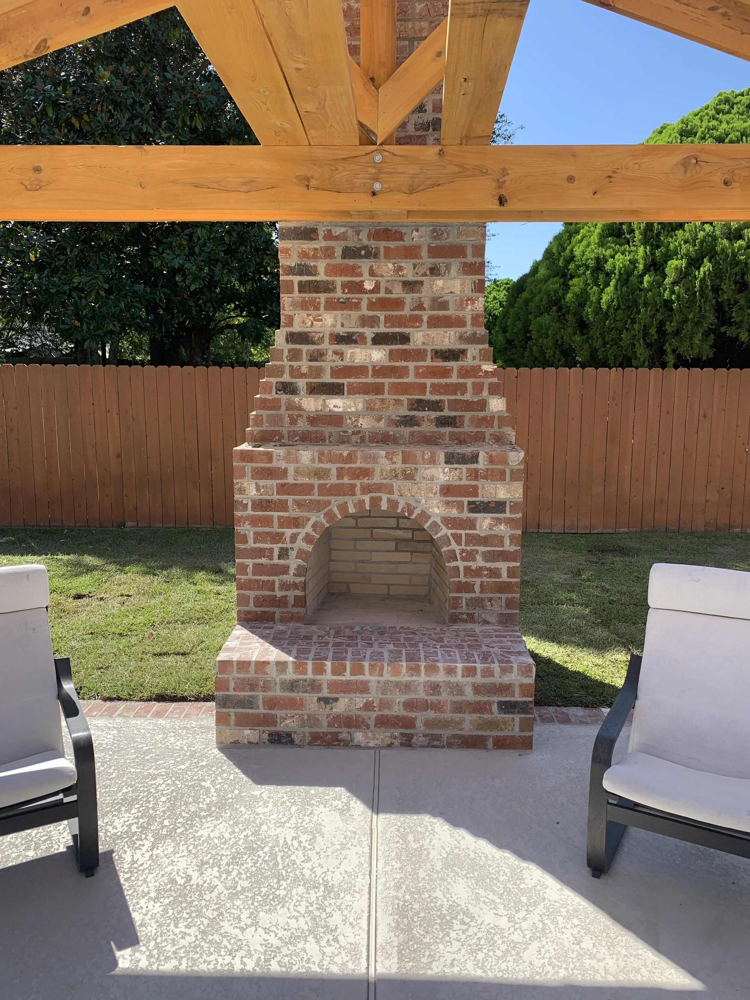 View of a backyard with a brick outdoor fireplace or oven, a wooden pergola, green trees, and patio chairs.