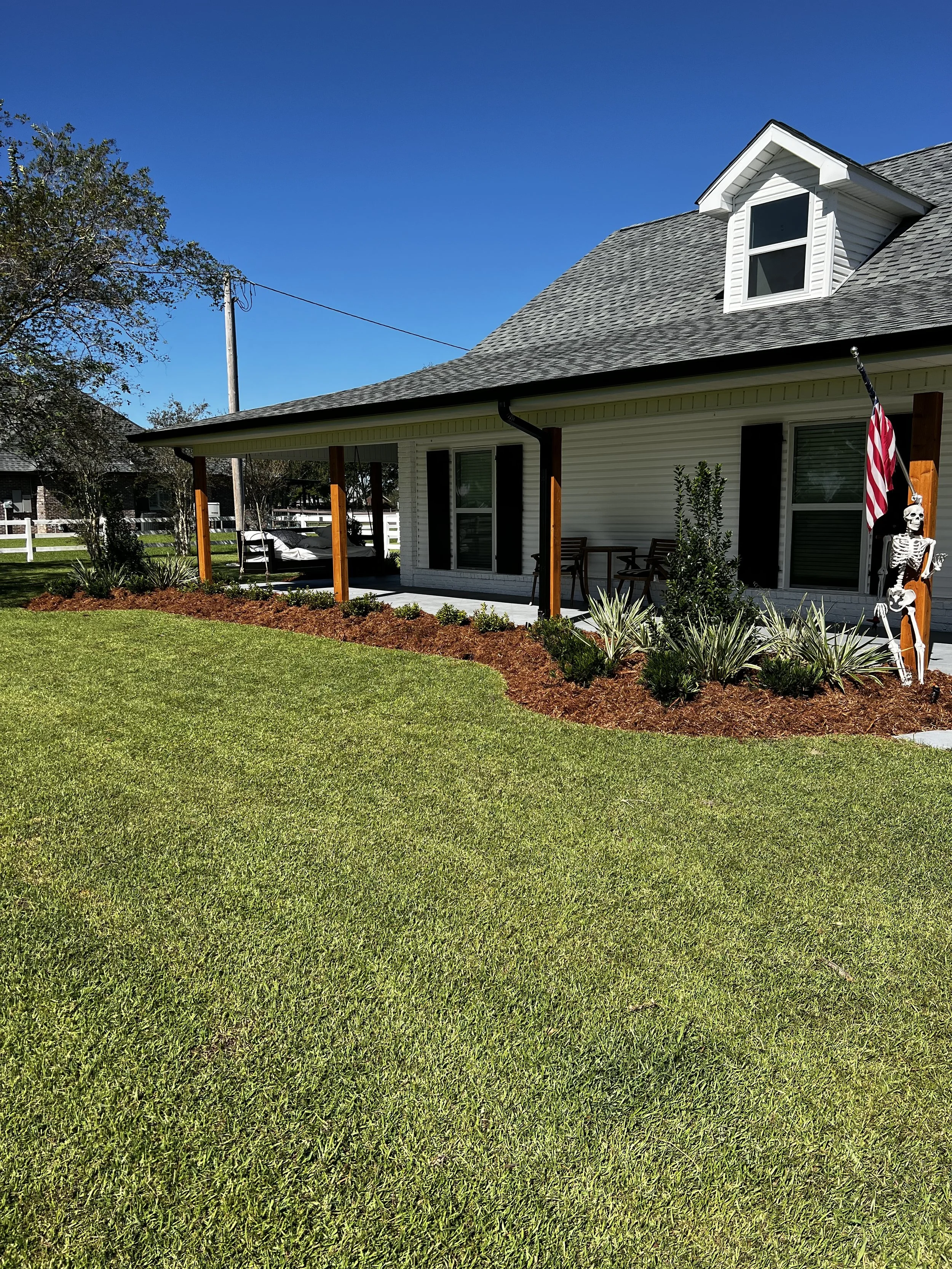 A front porch with a swing, potted plants, and a Halloween skeleton decoration holding a flag, attached to a white house with a gray roof and a dormer window, under a clear blue sky.