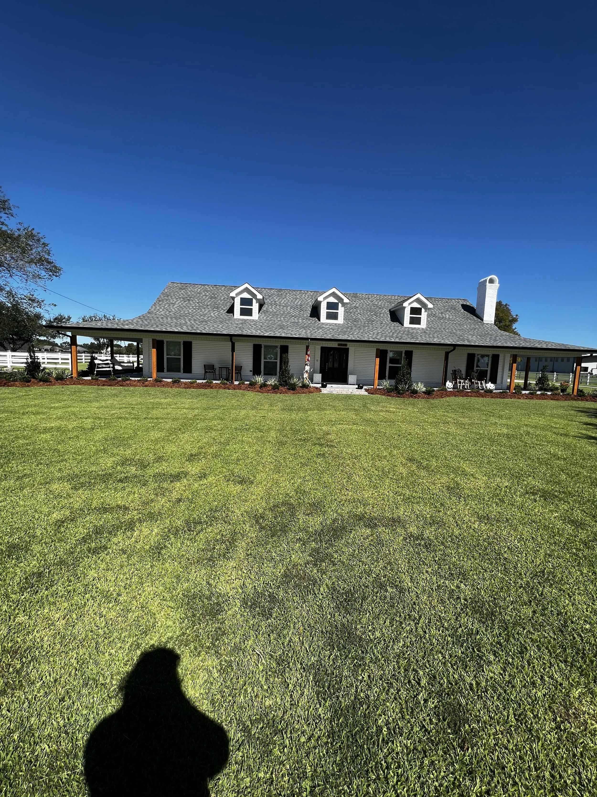 A large white house with a grey shingled roof and three dormer windows, surrounded by a well-maintained lawn and garden. The sky is clear and blue.