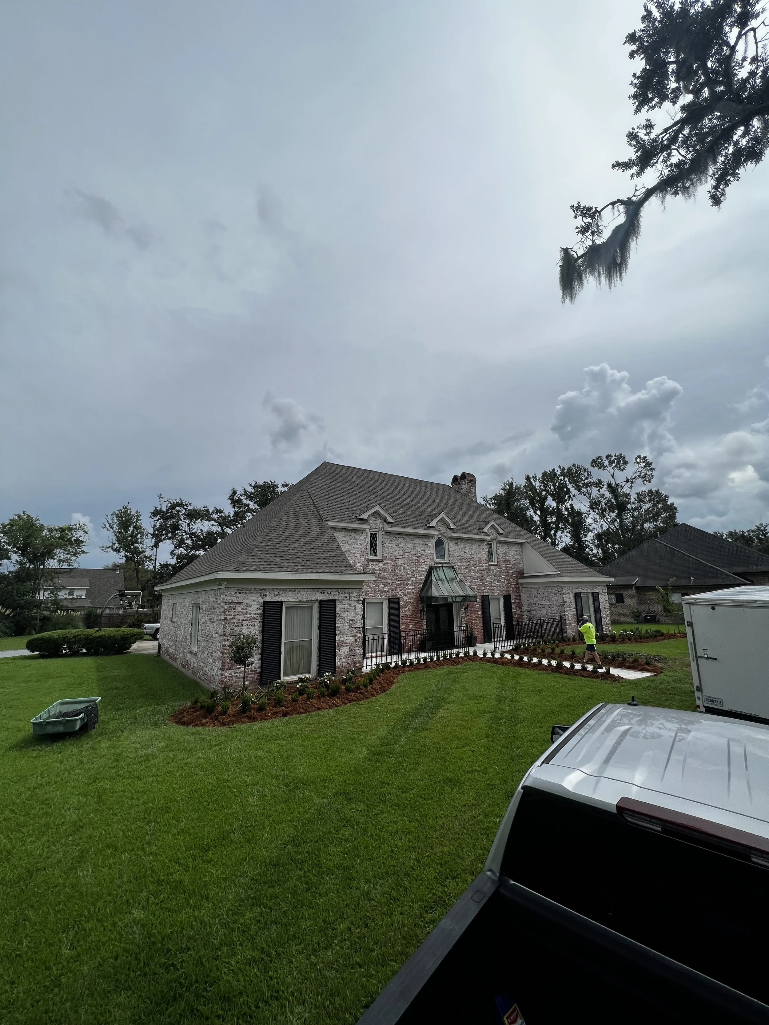 A house with brick walls and black shutters, with a newly landscaped front yard and a worker in a yellow shirt tending to the garden. The sky is partly cloudy, and a tree with moss-draped branches is visible on the right.