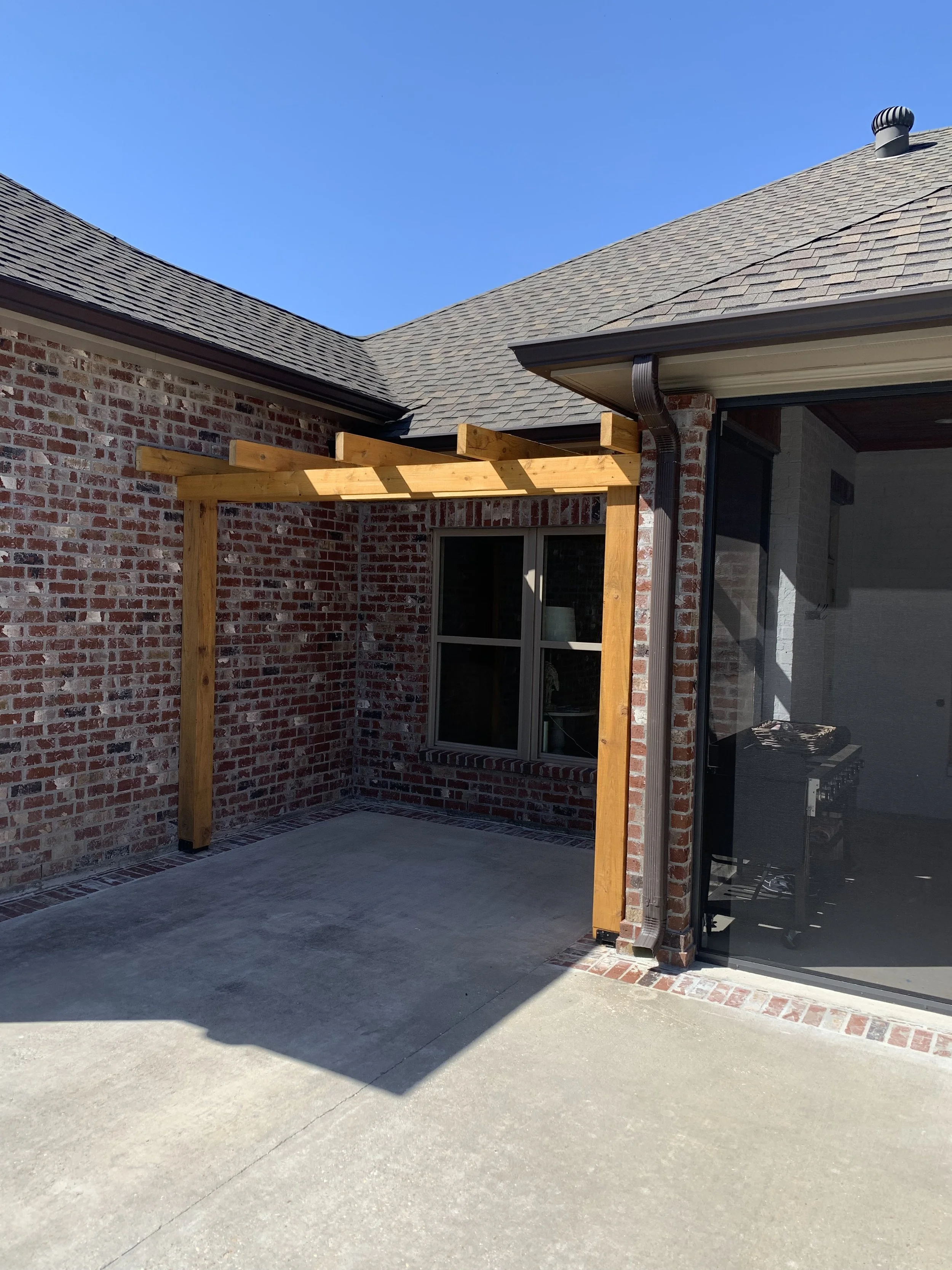 Backyard patio area with a brick house, wooden pergola under construction, sliding glass door, and a concrete floor.