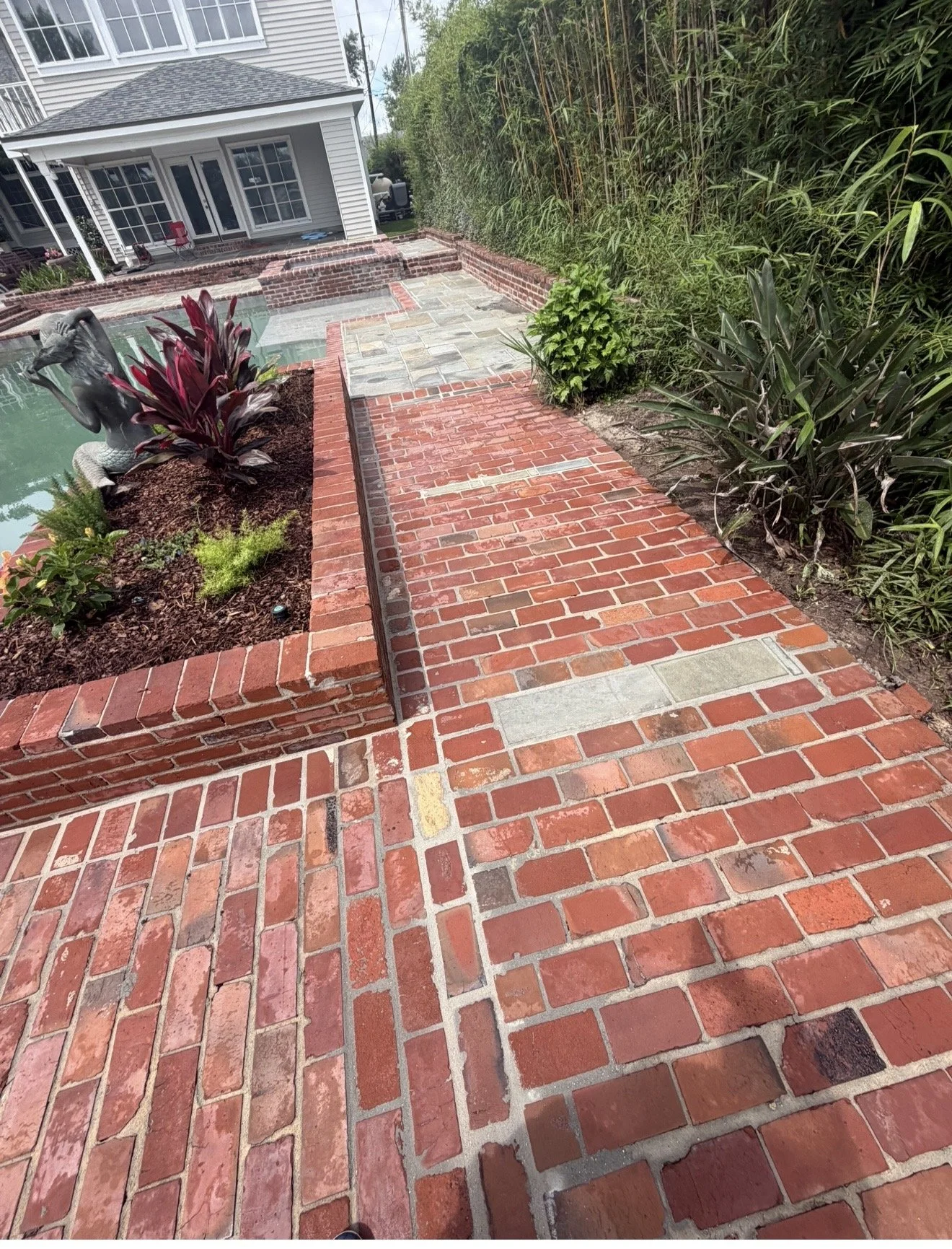 Backyard patio with brick pathway, brick raised garden bed with plants, a small fountain with a mermaid statue, and a house with a porch in the background.