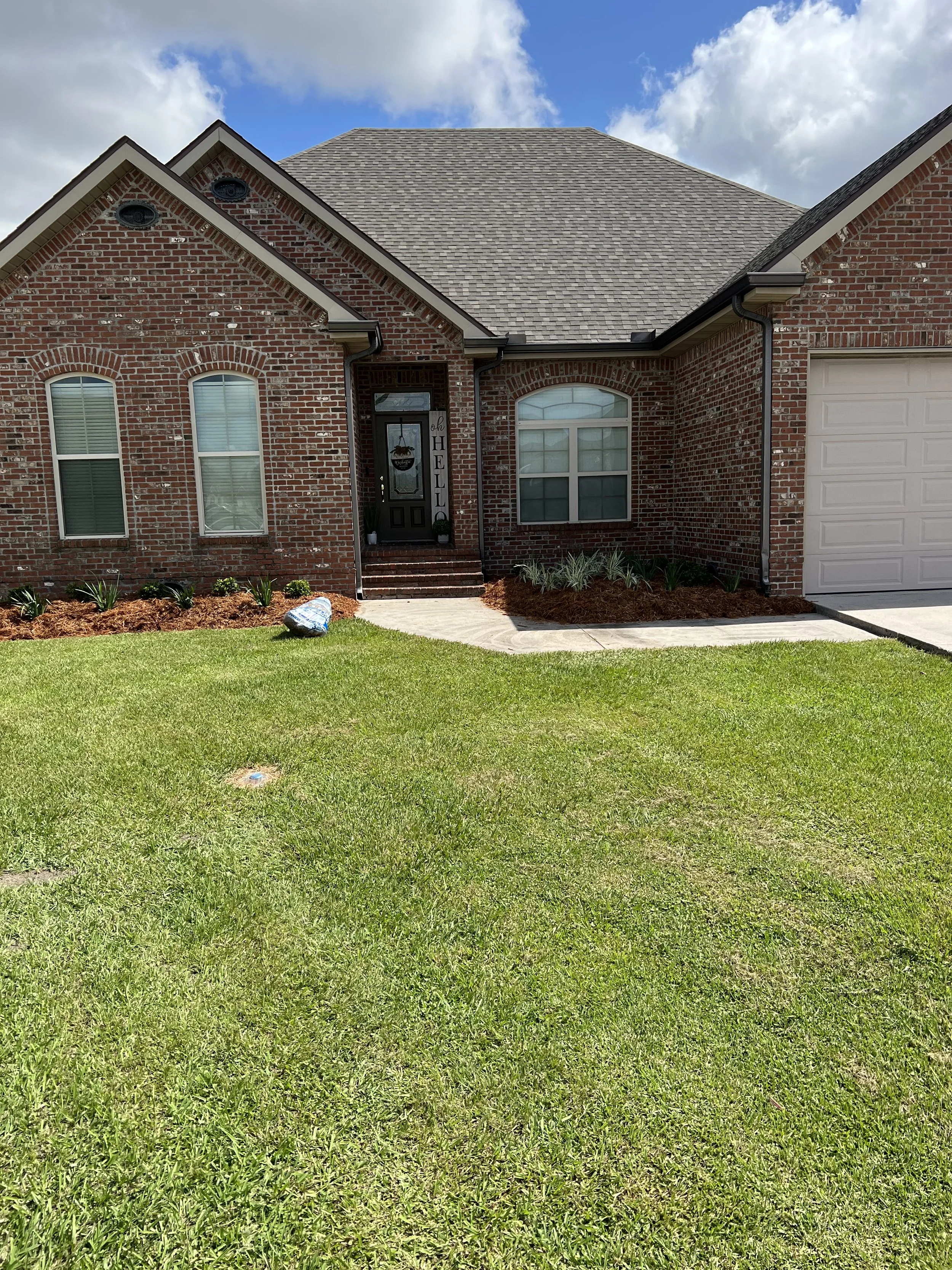 Front view of a brick house with a green lawn, flower beds, and a walkway leading to the front door under a partly cloudy sky.