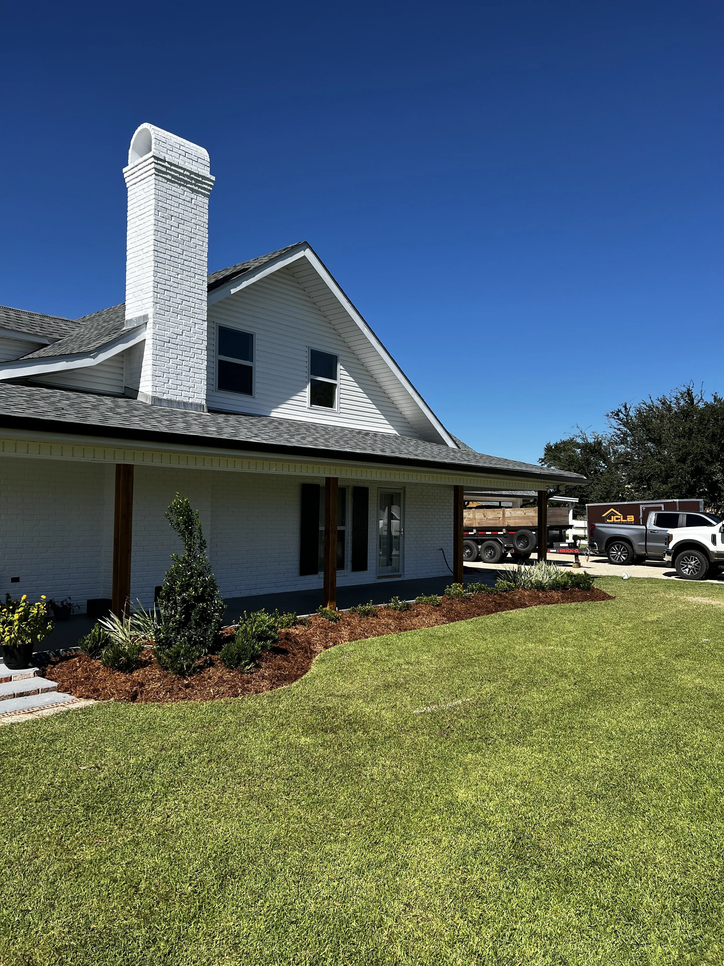 A house with white siding, a white brick chimney, new landscaping with plants and mulch, a well-maintained green lawn, and vehicles parked in the driveway under a clear blue sky.