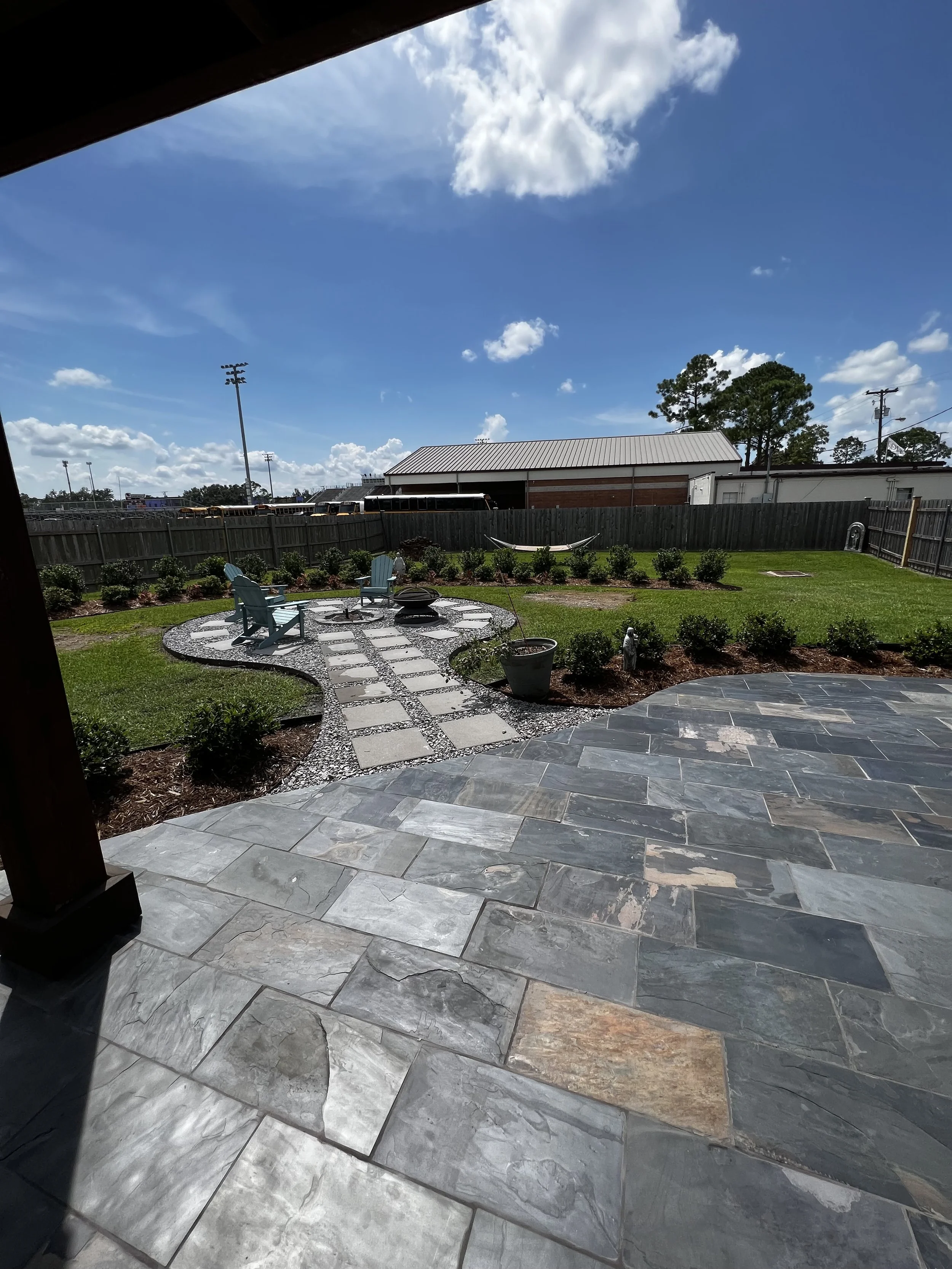 A backyard patio with stone flooring, garden area with bushes, Adirondack chairs around a fire pit, and a view of a blue sky with clouds, fence, and some trees.