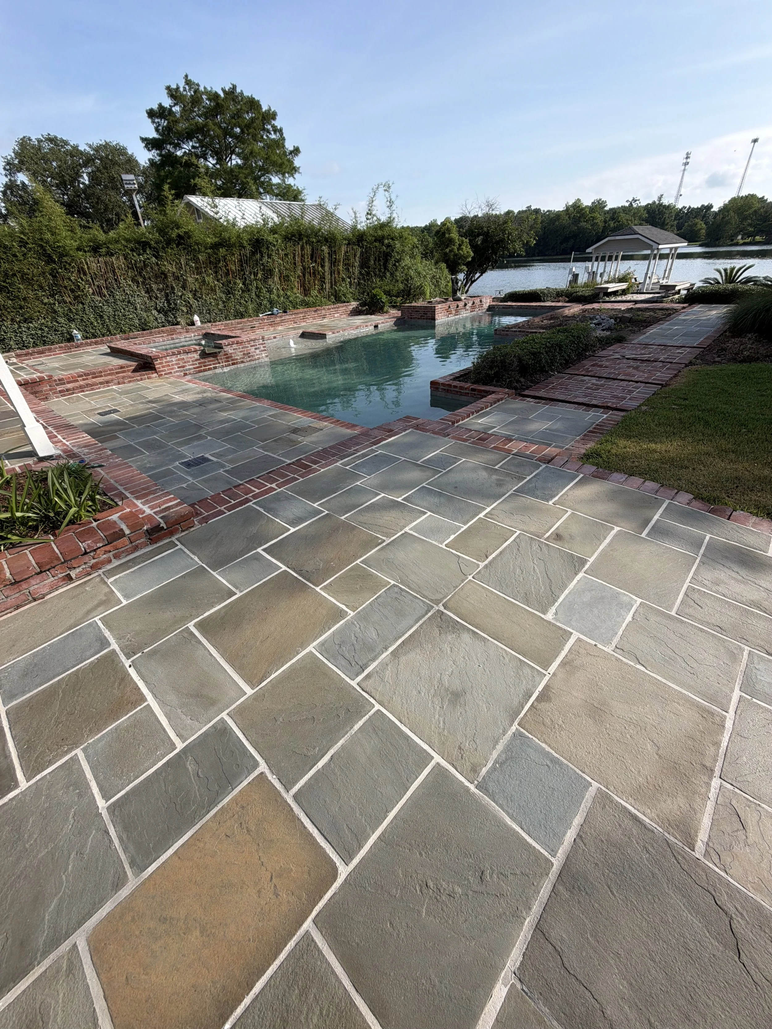 View of a backyard with a swimming pool, multi-colored stone tiles, brick accents, a brick wall, a gazebo over the water, benches, trees, and a lake in the background under a partly cloudy sky.