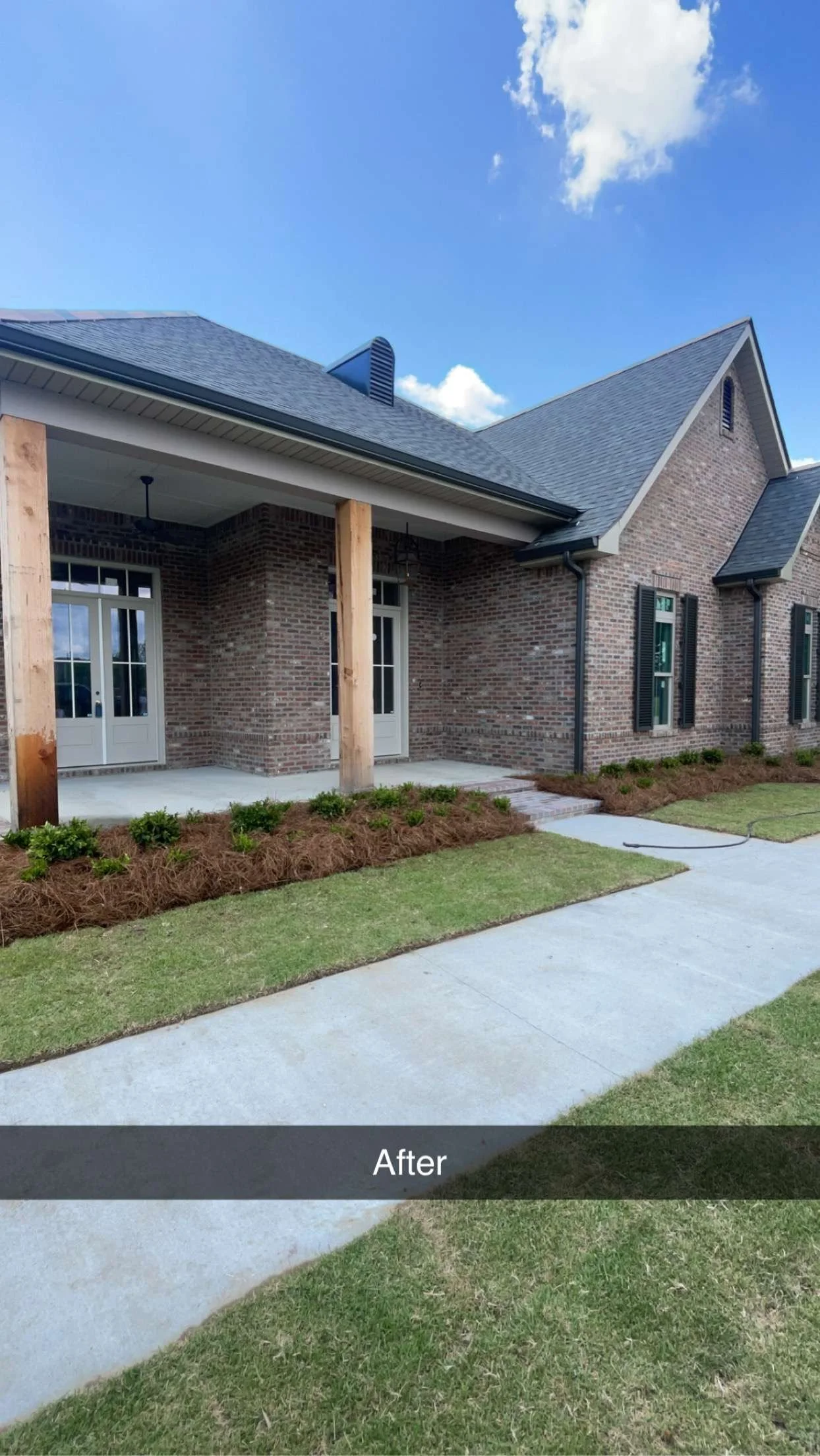 A newly renovated brick house with a covered patio area supported by wooden posts, double glass doors, black shutters, and a well-maintained lawn with small bushes. The sky is clear with a few clouds.