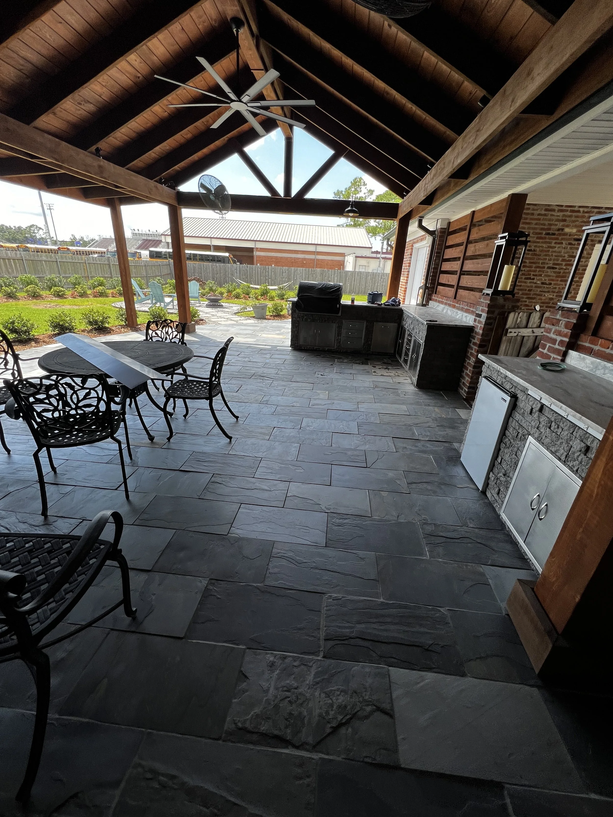 Covered outdoor patio area with a high wooden ceiling, ceiling fan, outdoor kitchen with a grill, and a patio table with chairs, overlooking a garden with plants and a lawn.