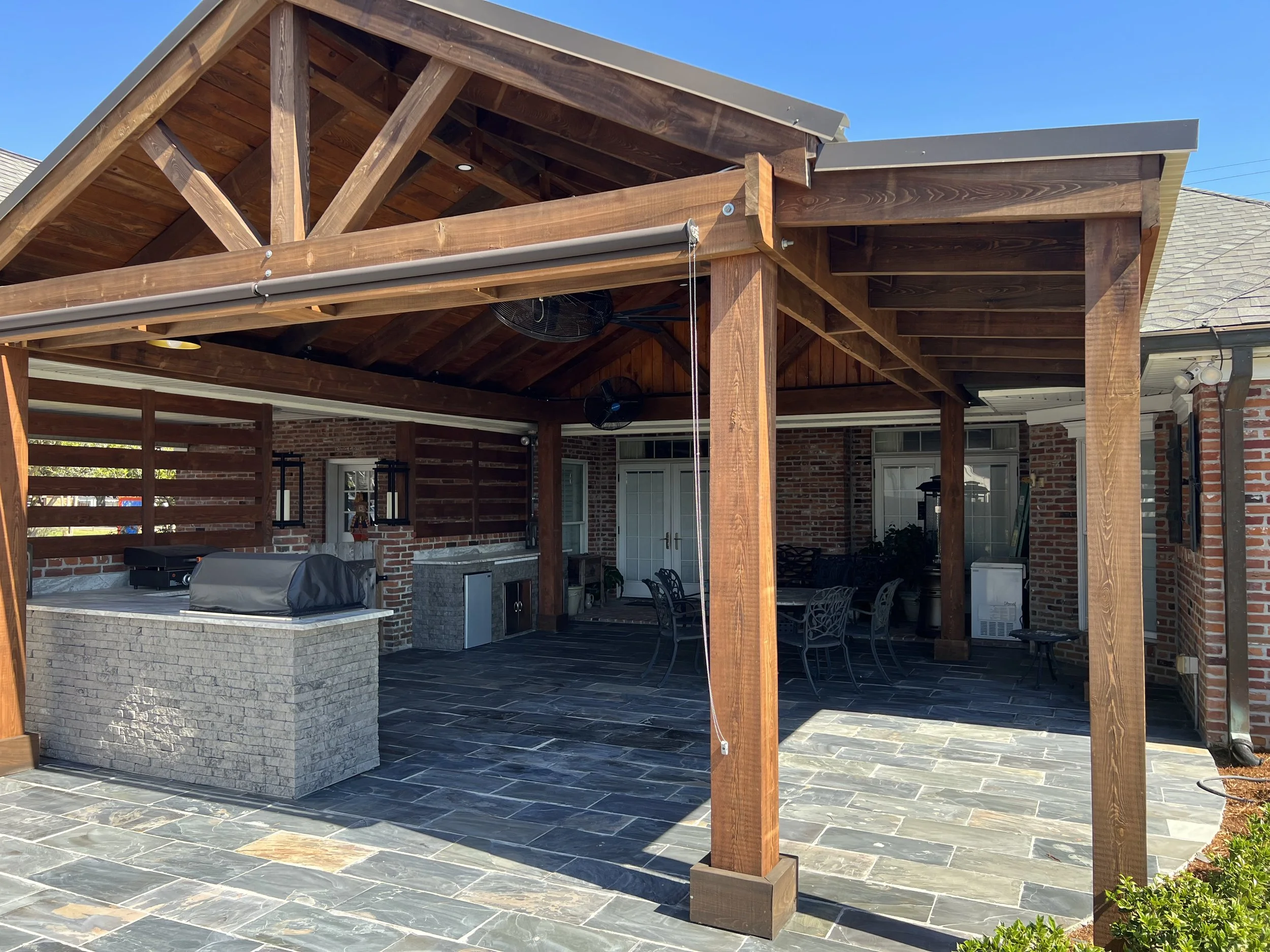 A backyard patio area with a wooden pergola structure, brick fireplace, and outdoor seating. The space features a stone or ceramic tiled floor and is under a blue sky.