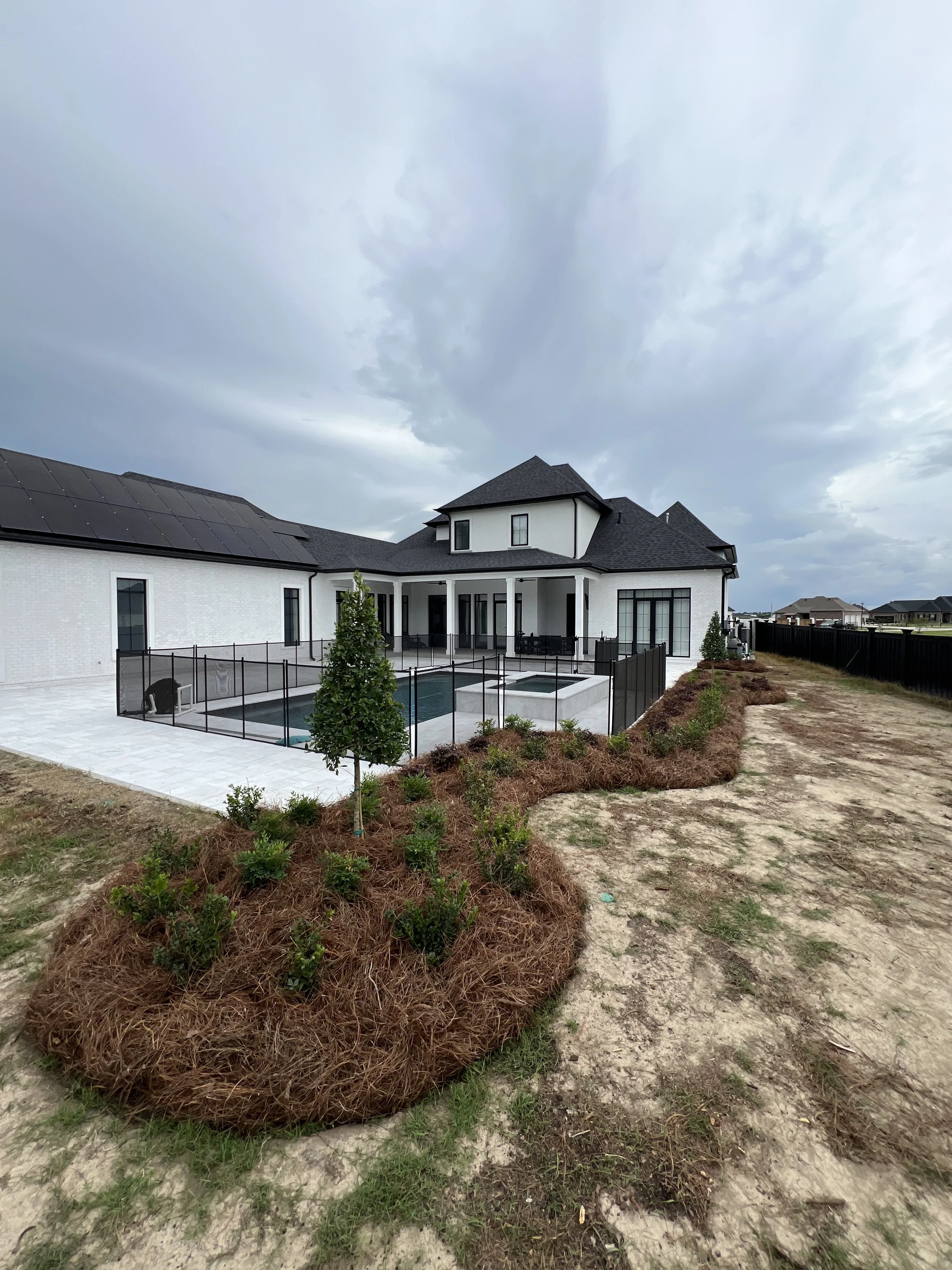 A large white house with black roof shingles and solar panels on one side, a backyard patio with a pool surrounded by a black fence, some small trees, and an overcast sky with dark clouds.