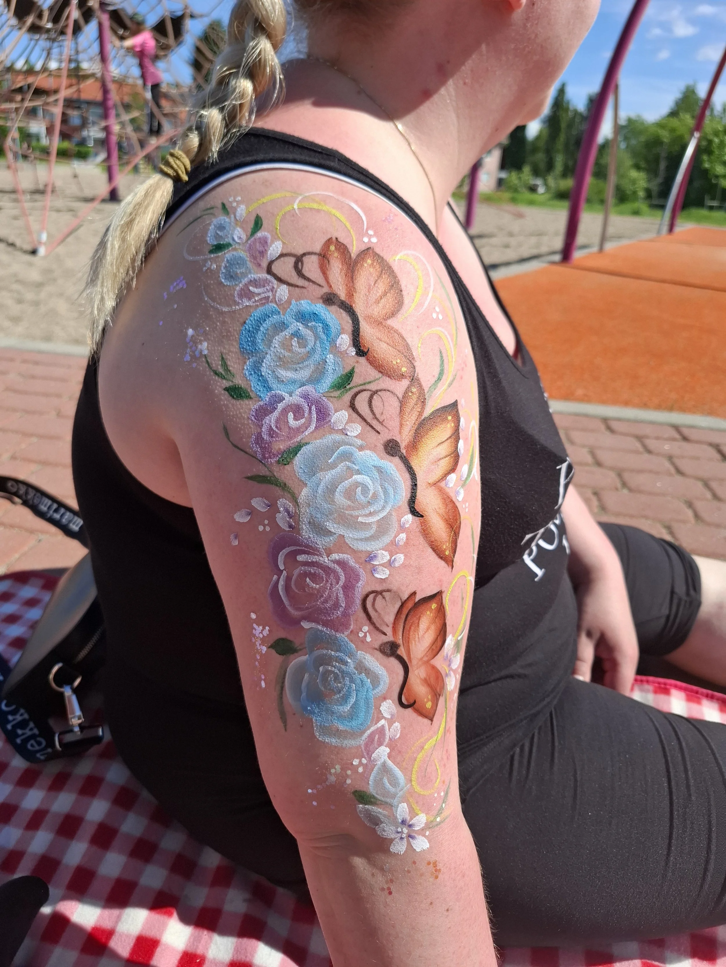 Naisen oikean olkapään alueella on värikäs ihomaalaus, jossa on kukkia ja perhonen, sekä suihkemaista glitteriä ja värikkäitä koristeita.