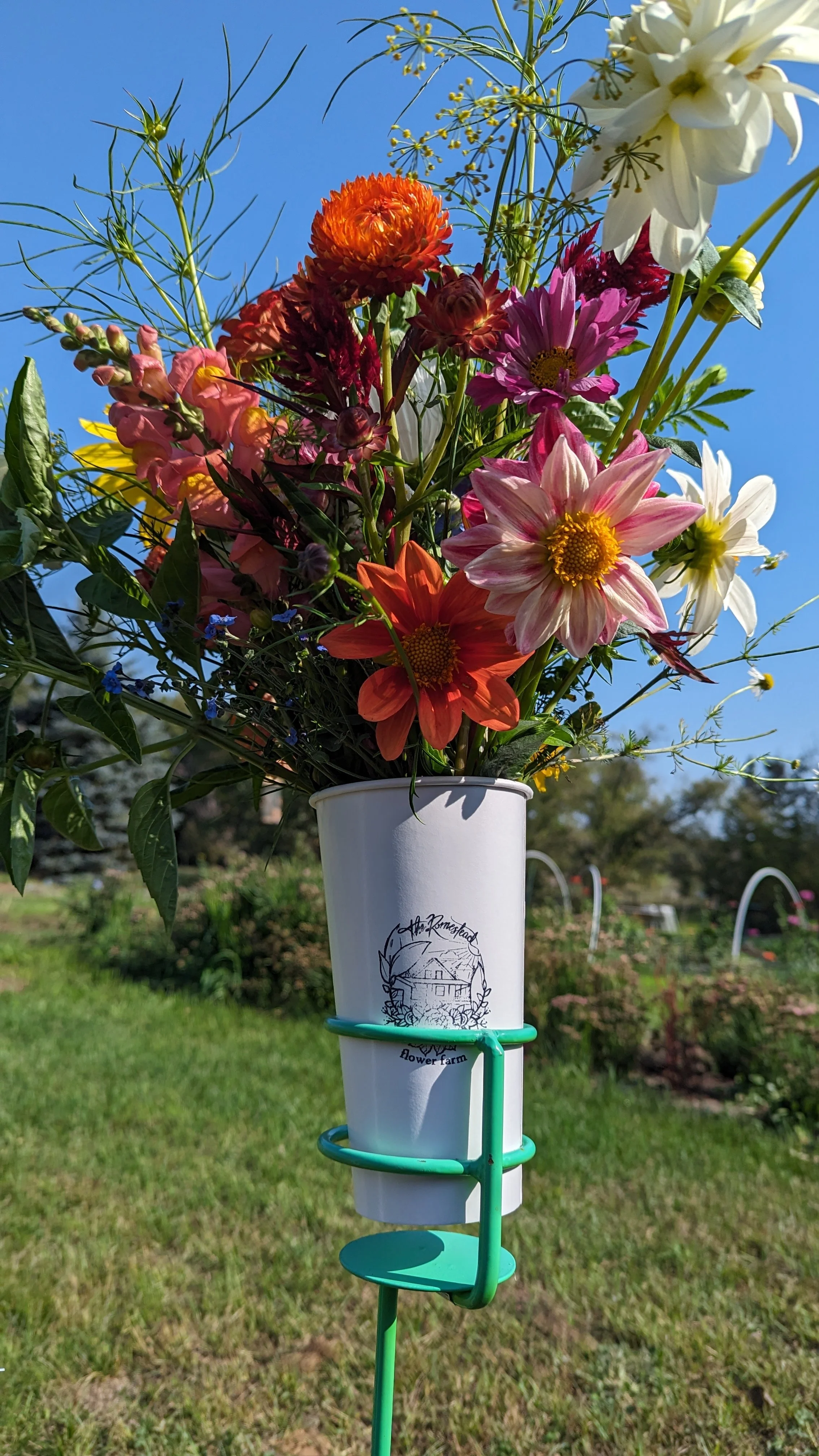 A white flower farm cup with a floral arrangement of various colorful flowers, including white lilies, pink zinnias, orange and red dahlias, and purple aster, is fastened to a green metal stake outdoors against a backdrop of blue sky and greenery.   