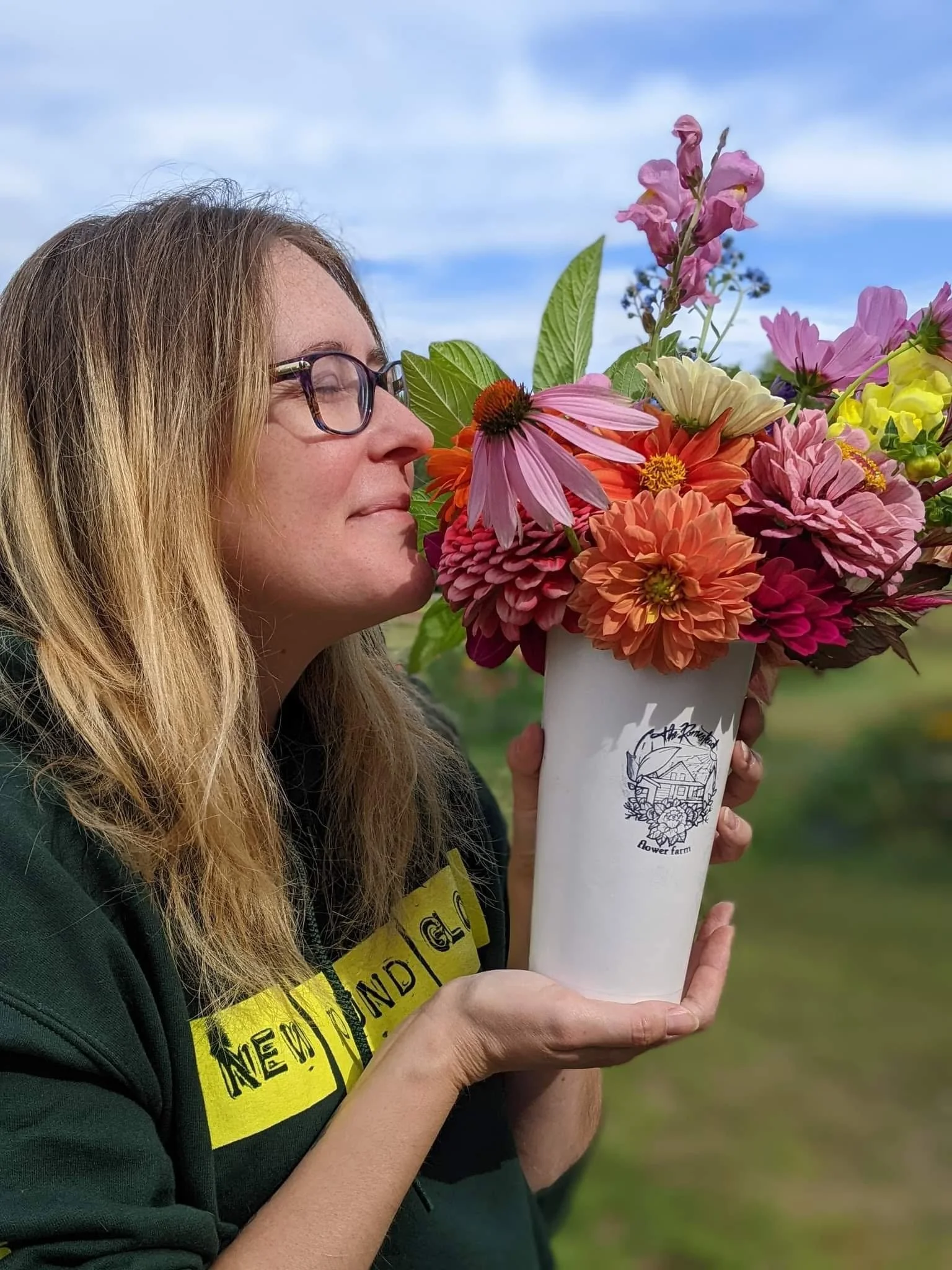Woman with glasses smelling a colorful bouquet of flowers in a white cup outdoors against a blue sky.