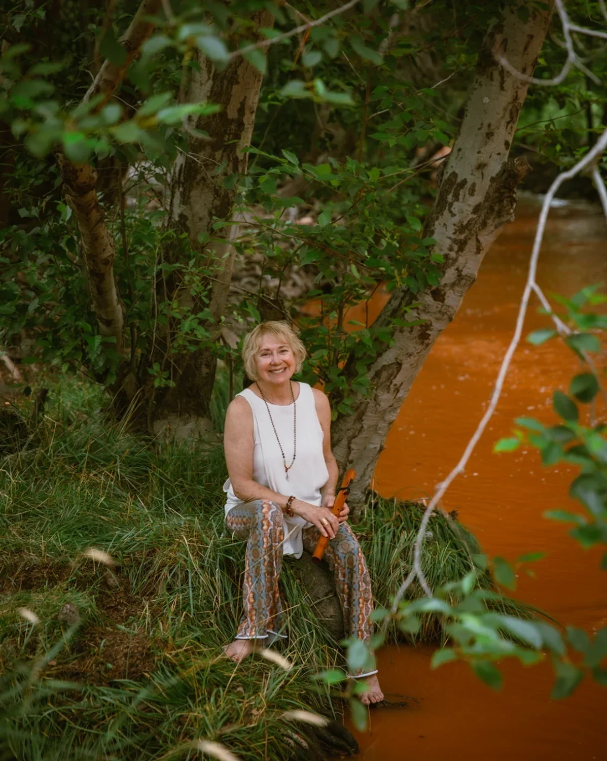 A smiling elderly woman sitting on a grassy bank by a river, holding a flute, surrounded by trees and greenery.