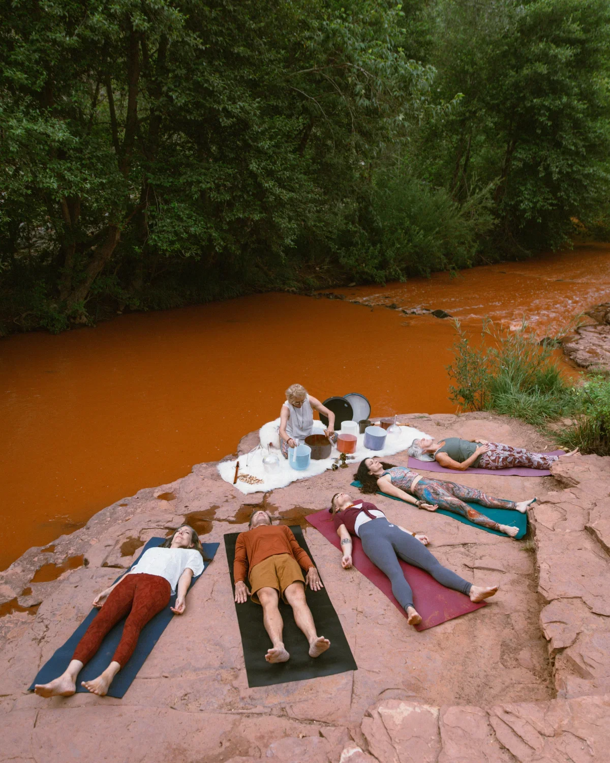 Group of people practicing yoga by a river on a rocky outcrop surrounded by trees.
